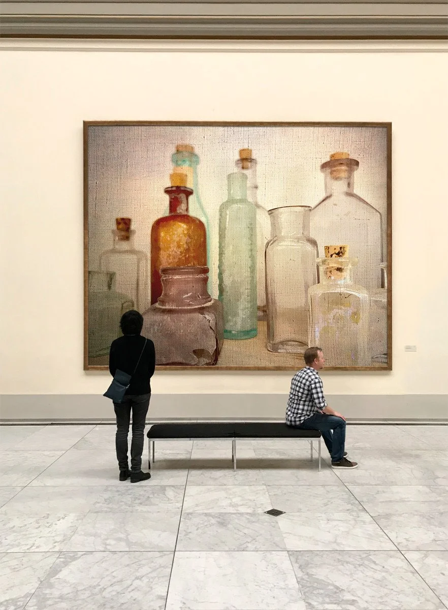 People sitting and standing in front of a large painting of glass bottles in an art gallery.