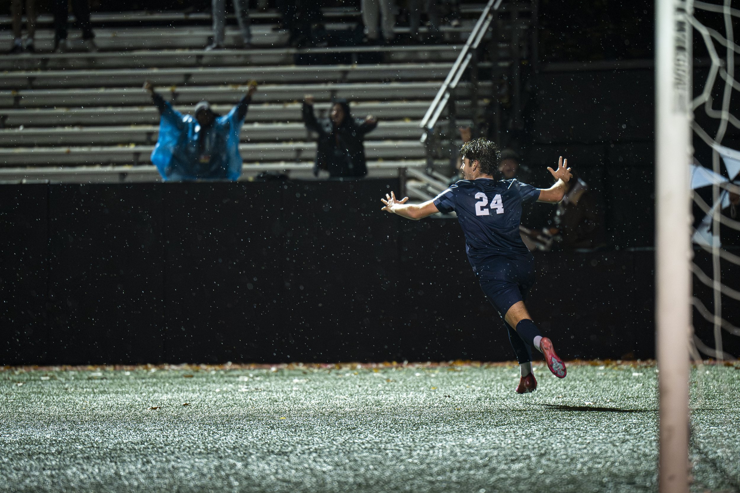 A soccer player wearing a navy blue jersey with the number 24 is celebrating after scoring a goal, running towards the corner flag on a rain-soaked pitch, with spectators in the stands in the background.