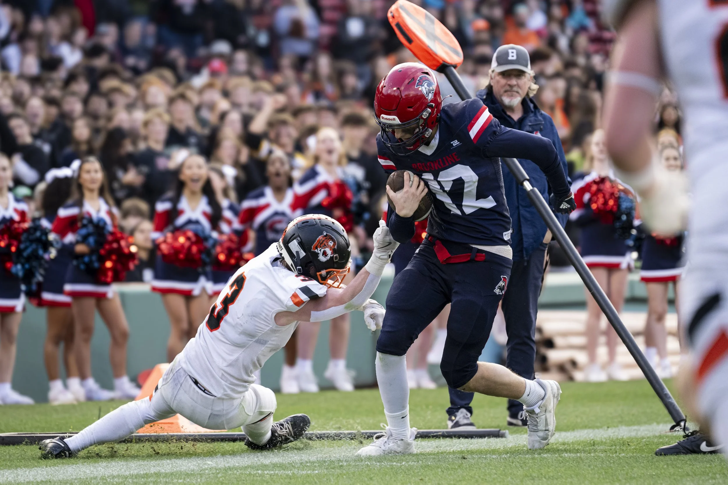 American football game showing a player in a blue jersey with the number 12 running with the football, being tackled by a player in a white jersey with the number 3, with cheerleaders and crowd in the background.
