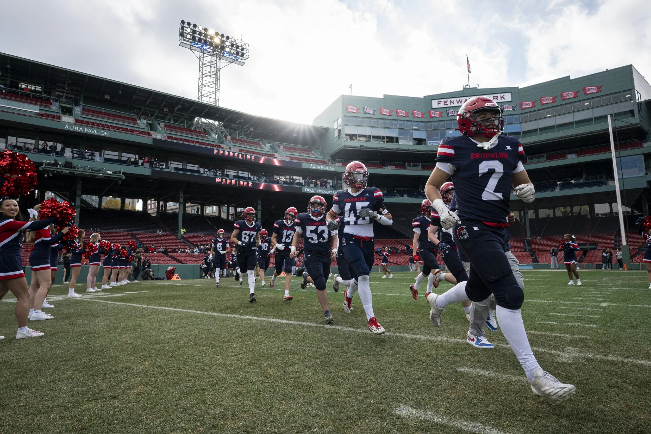 American football players in dark blue and red uniforms running onto the field at Fenway Park, with cheerleaders on the sidelines and a sunny sky overhead.