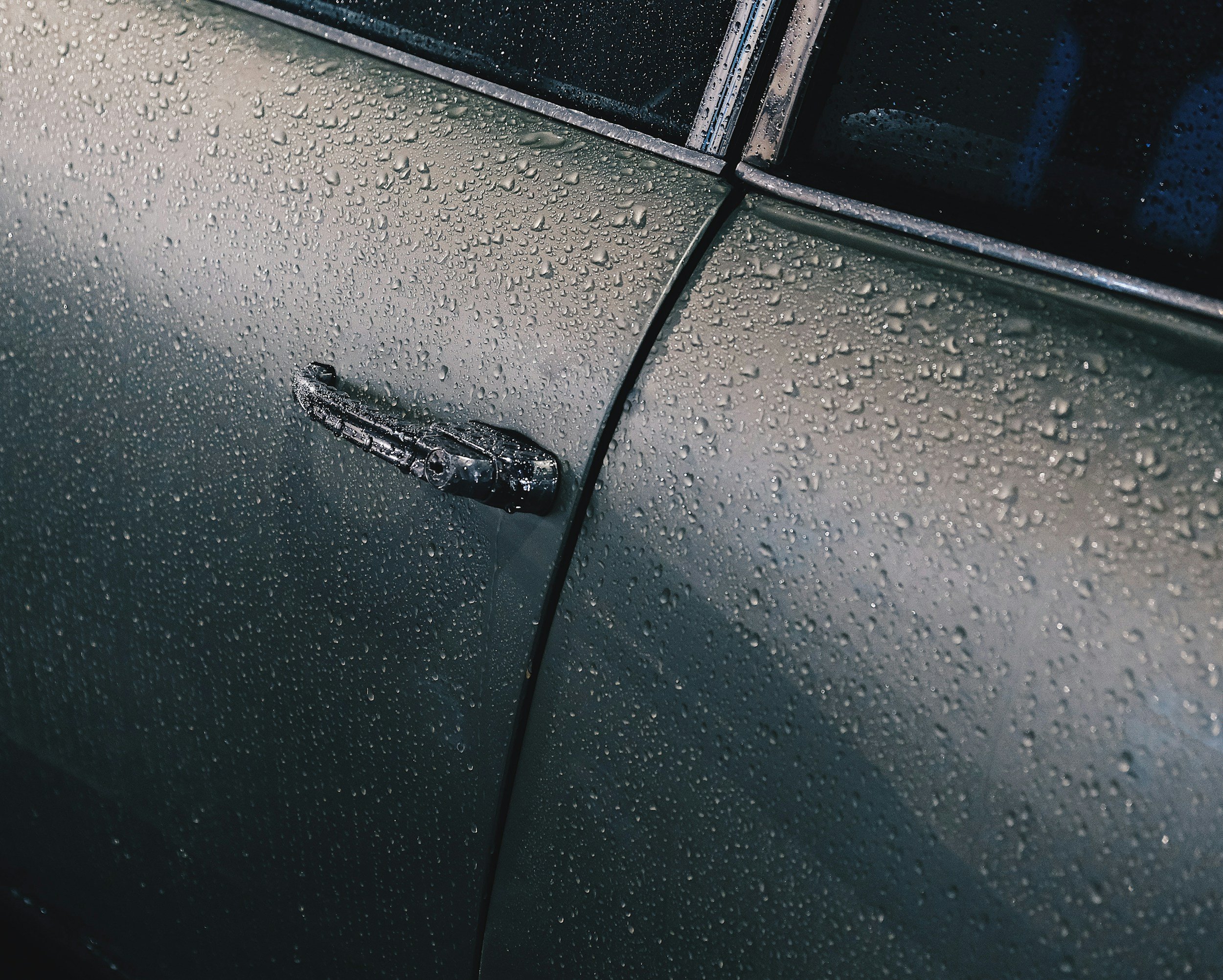 Close-up of a silver car's door handle covered in water droplets, with a wet surface and raindrops on the car body.