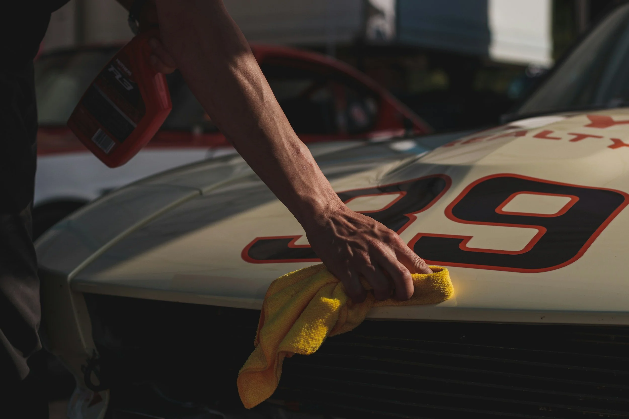 Person cleaning the hood of a race car with a yellow cloth while holding a spray bottle.