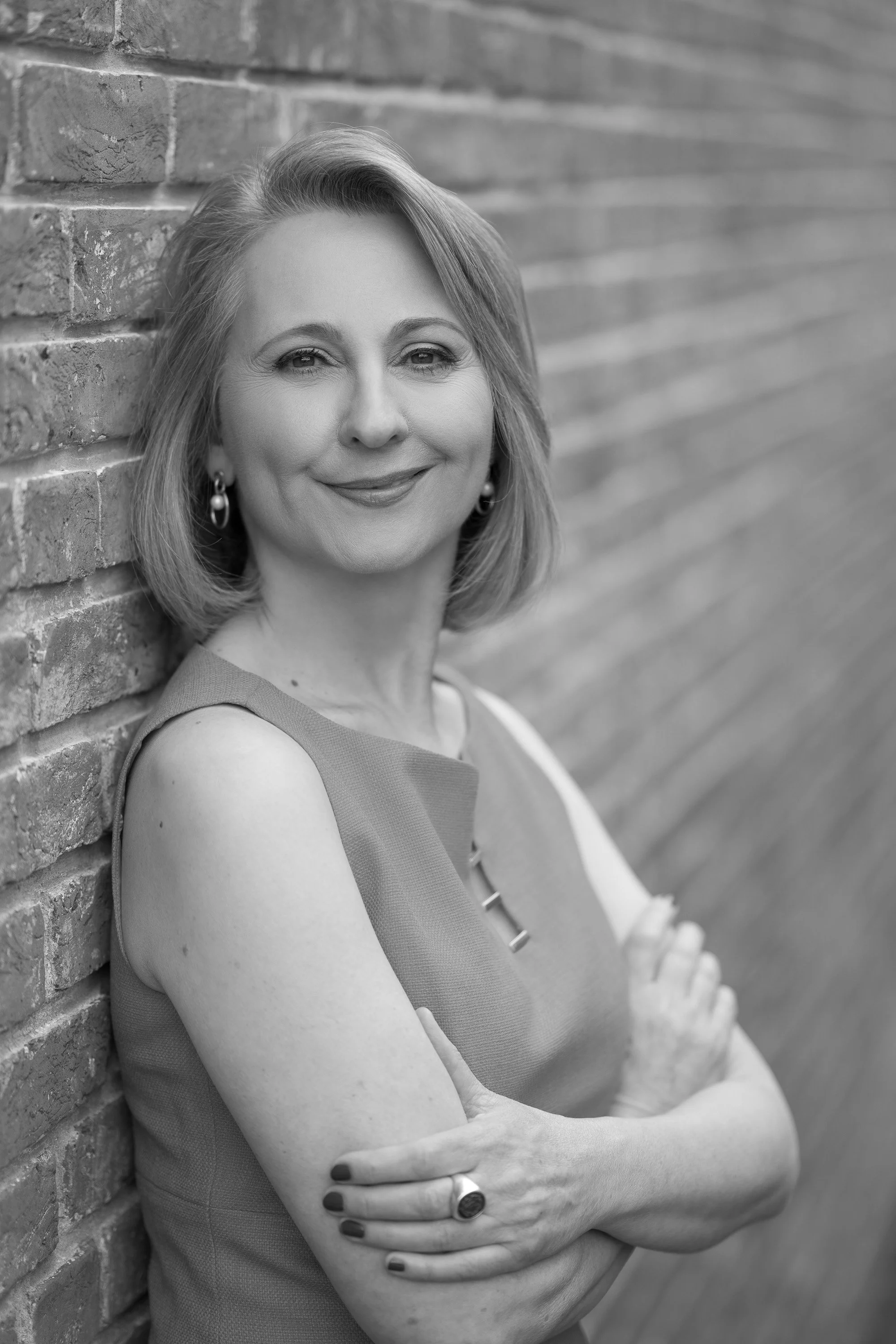 Black and white portrait of a middle-aged woman with shoulder-length hair, smiling, standing against a brick wall with crossed arms.
