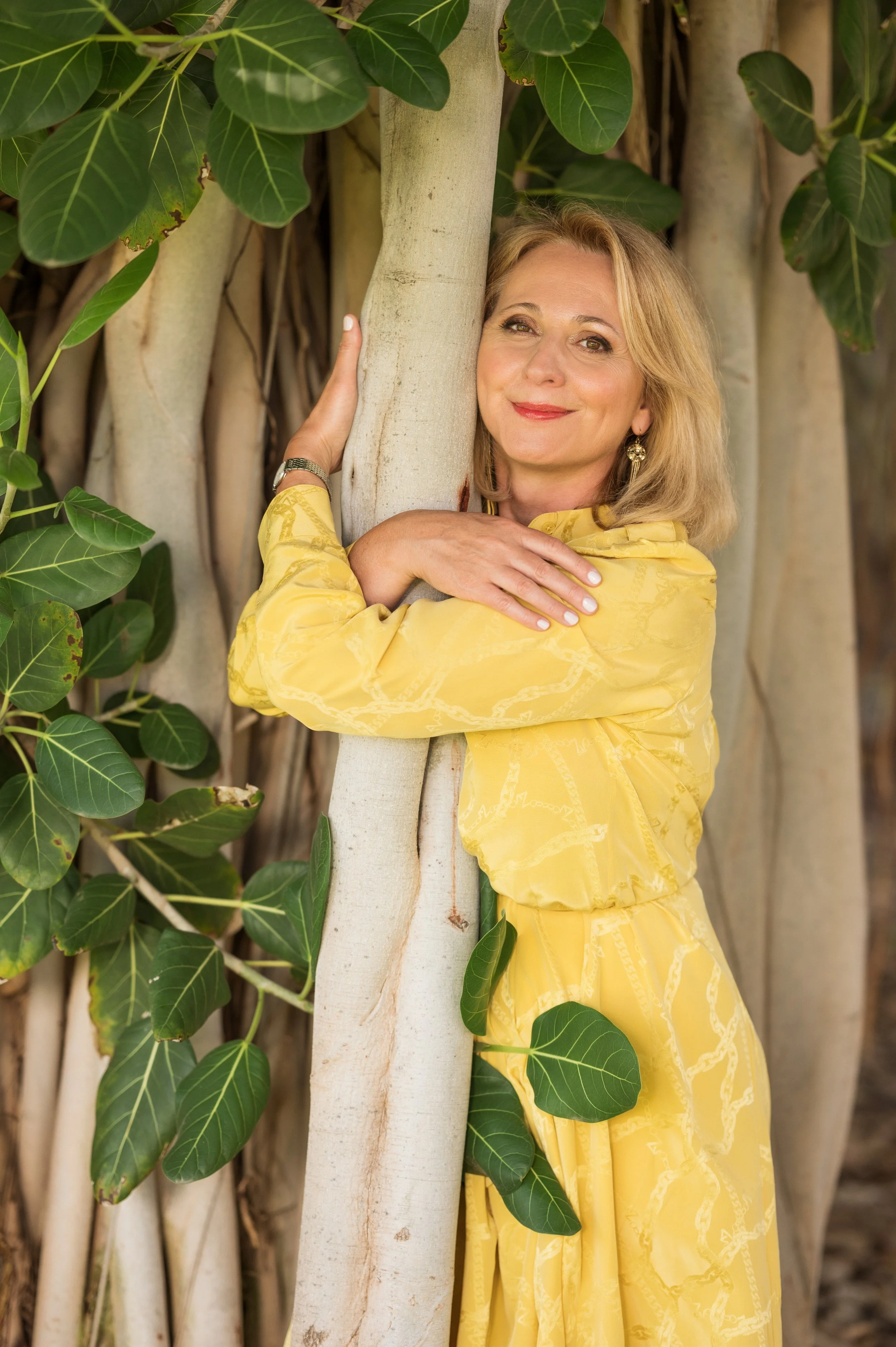 A woman in a yellow dress hugging a large tree with a background of green leaves and tree trunks