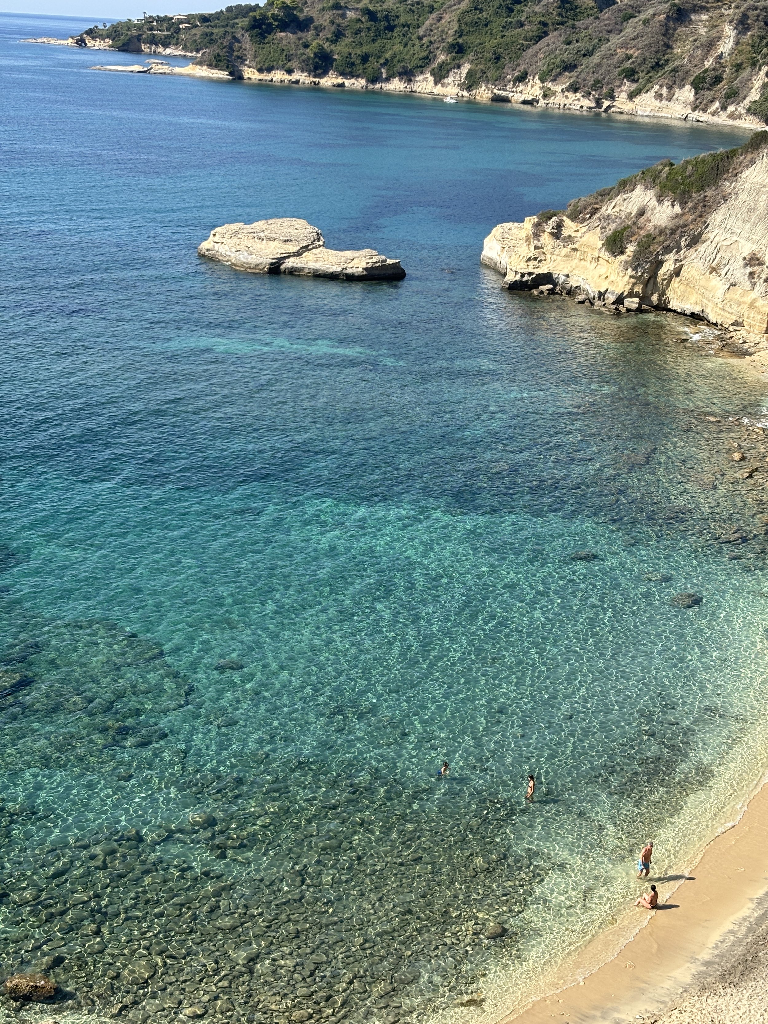 A clear, turquoise bay with rocky shoreline and a few people swimming and relaxing on the sand.