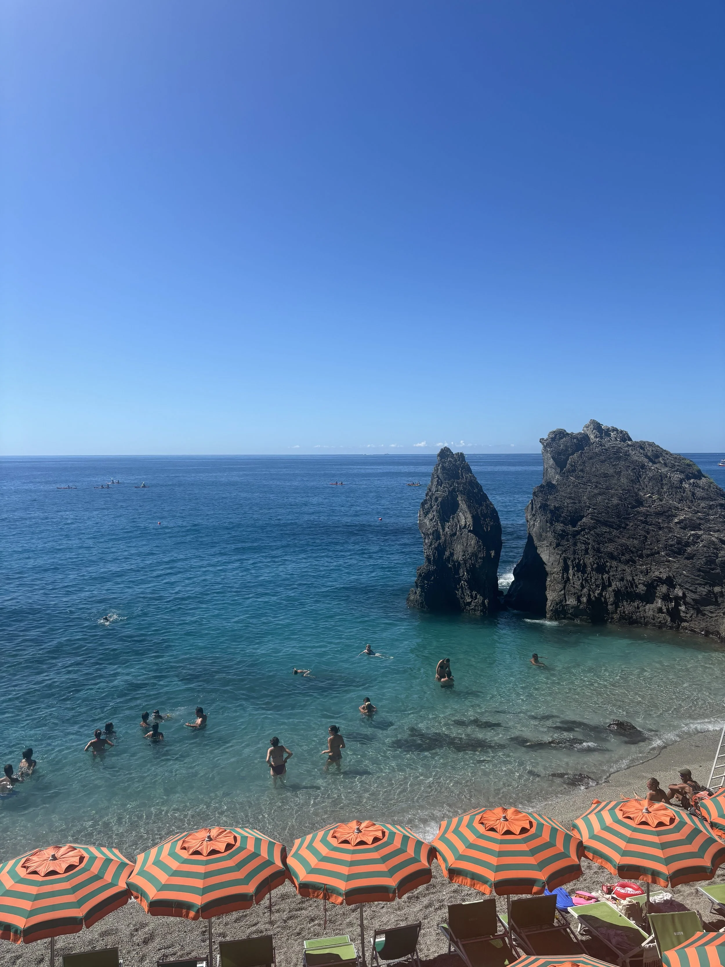 Beach with orange and green striped umbrellas, people swimming and relaxing by the shoreline, large rock formations, and a clear blue sky.