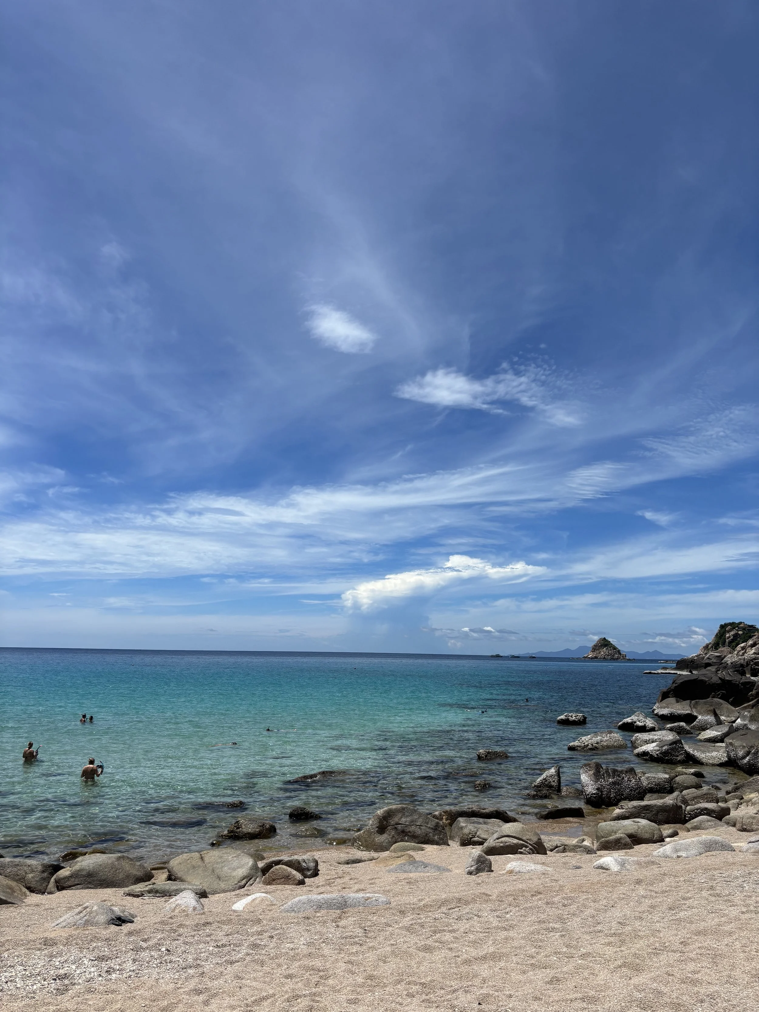 Beach with rocks and clear turquoise water, a few people swimming, and a partly cloudy sky.