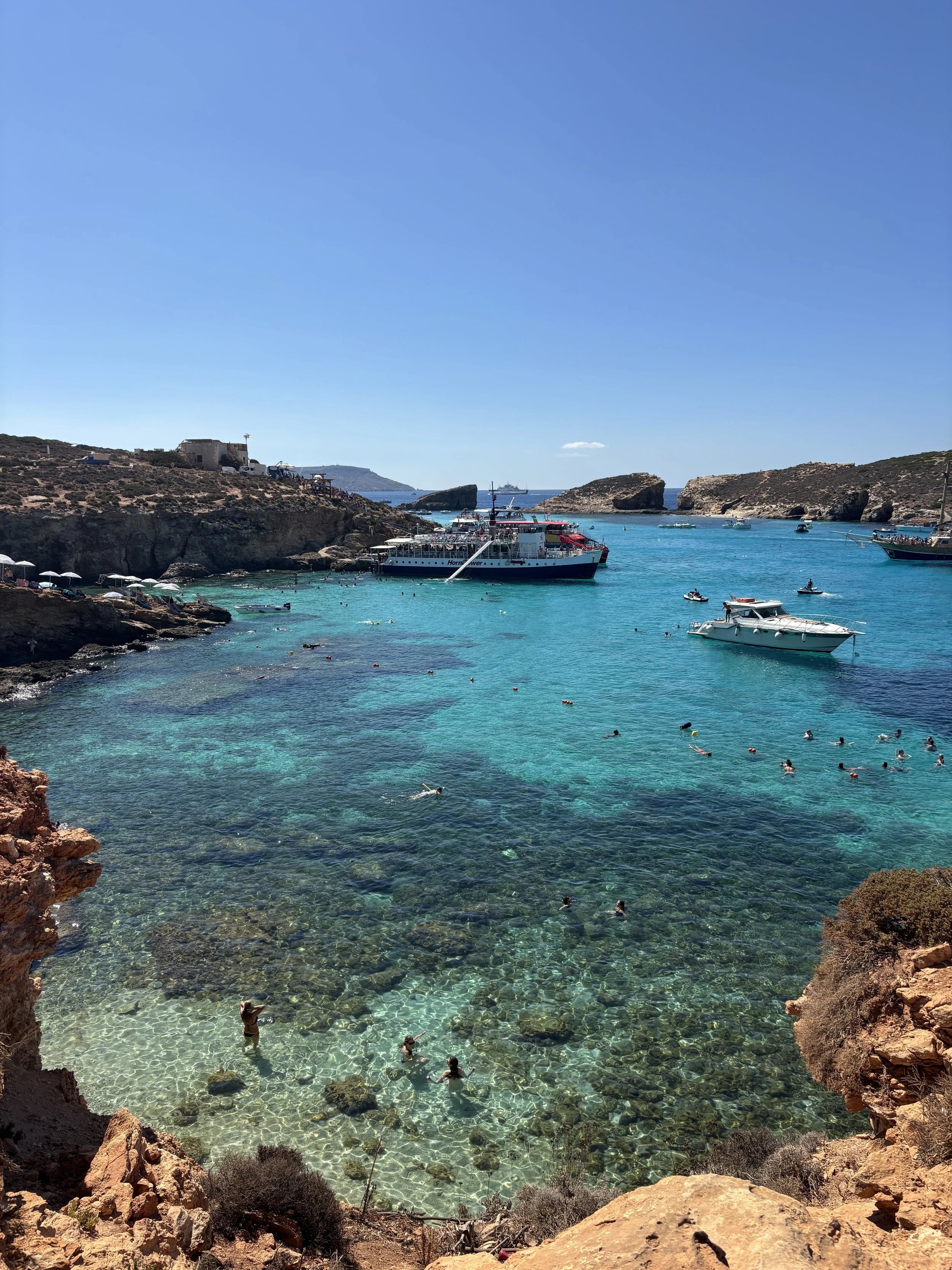 A clear blue bay with rocky shores, boats floating in the water, and people swimming and snorkeling in the turquoise water on a sunny day.