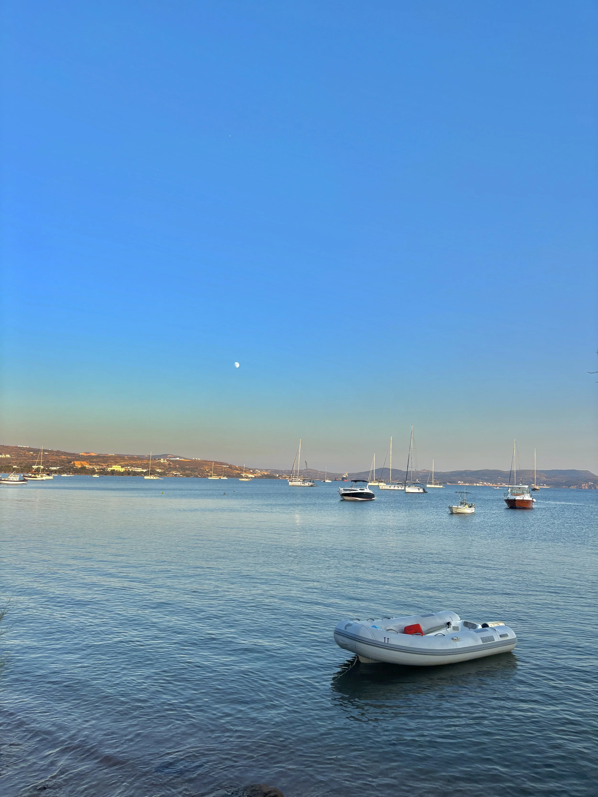 Calm harbor with sailboats and a small inflatable boat, distant shoreline, clear blue sky with visible moon, during daytime.