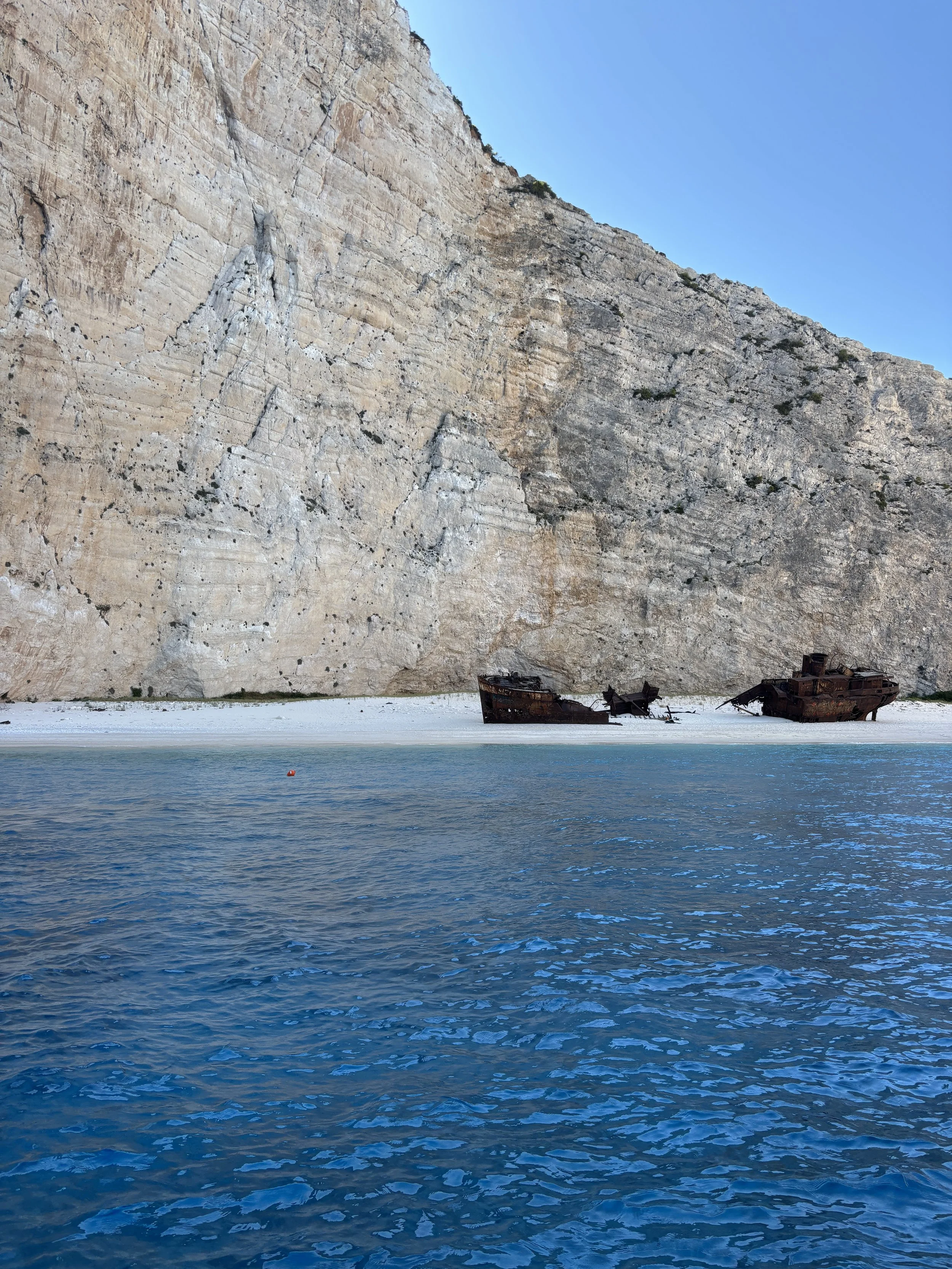 Shipwrecks on a sandy beach at the base of a towering cliff with a clear blue sky.