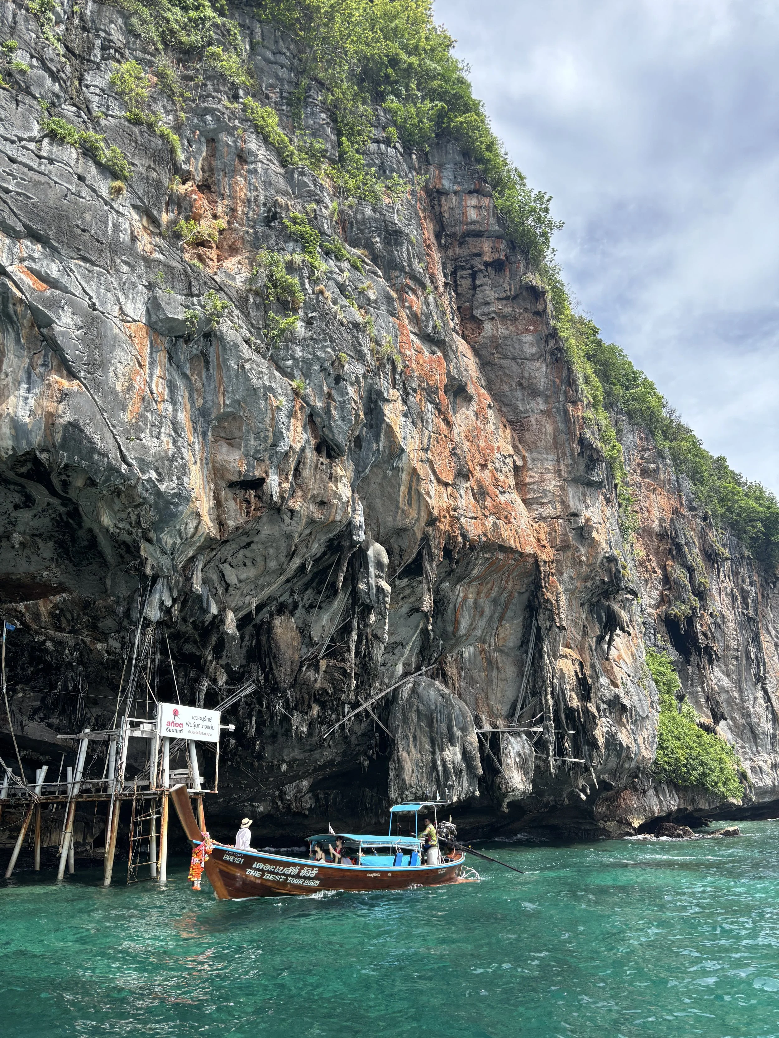 Boat docked near a rocky overhang with climbing ropes, turquoise water, and lush greenery on tall cliff.