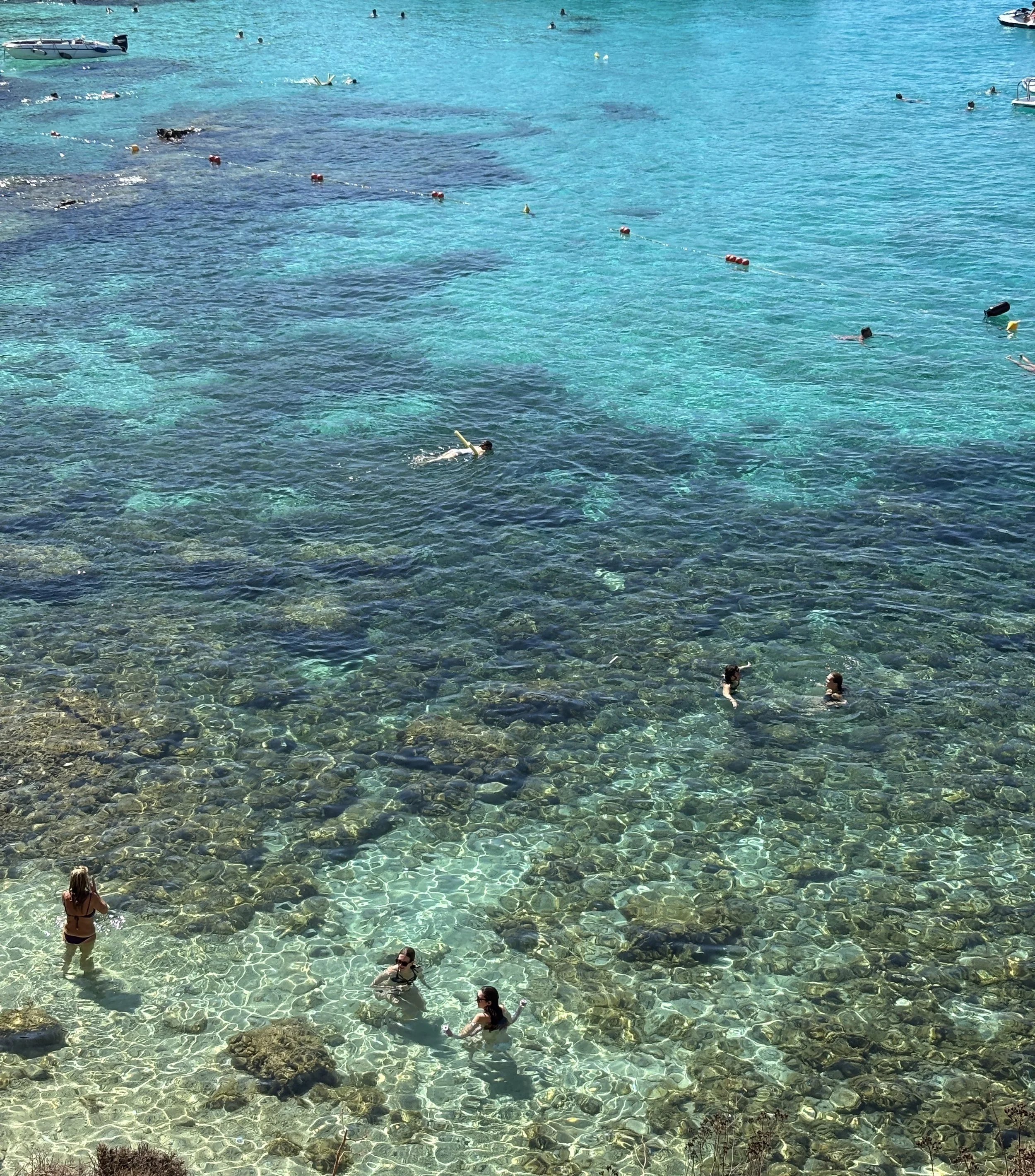 People swimming and wading in clear turquoise ocean water near rocky shoreline, with boats and floating buoys visible in the distance.