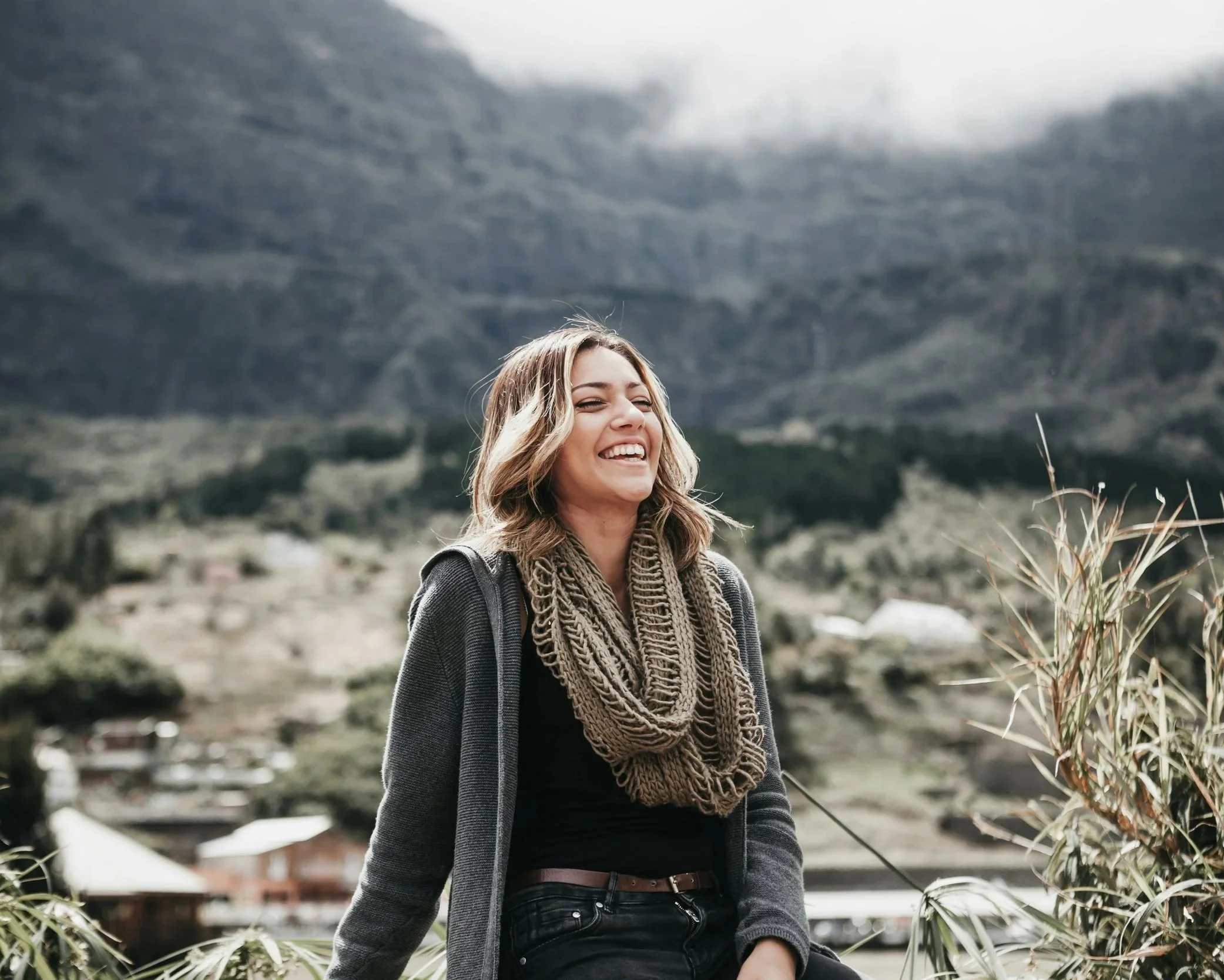 A woman with blonde wavy hair smiling outdoors in a mountainous area, wearing a dark jacket, a brown chunky scarf, and black jeans.