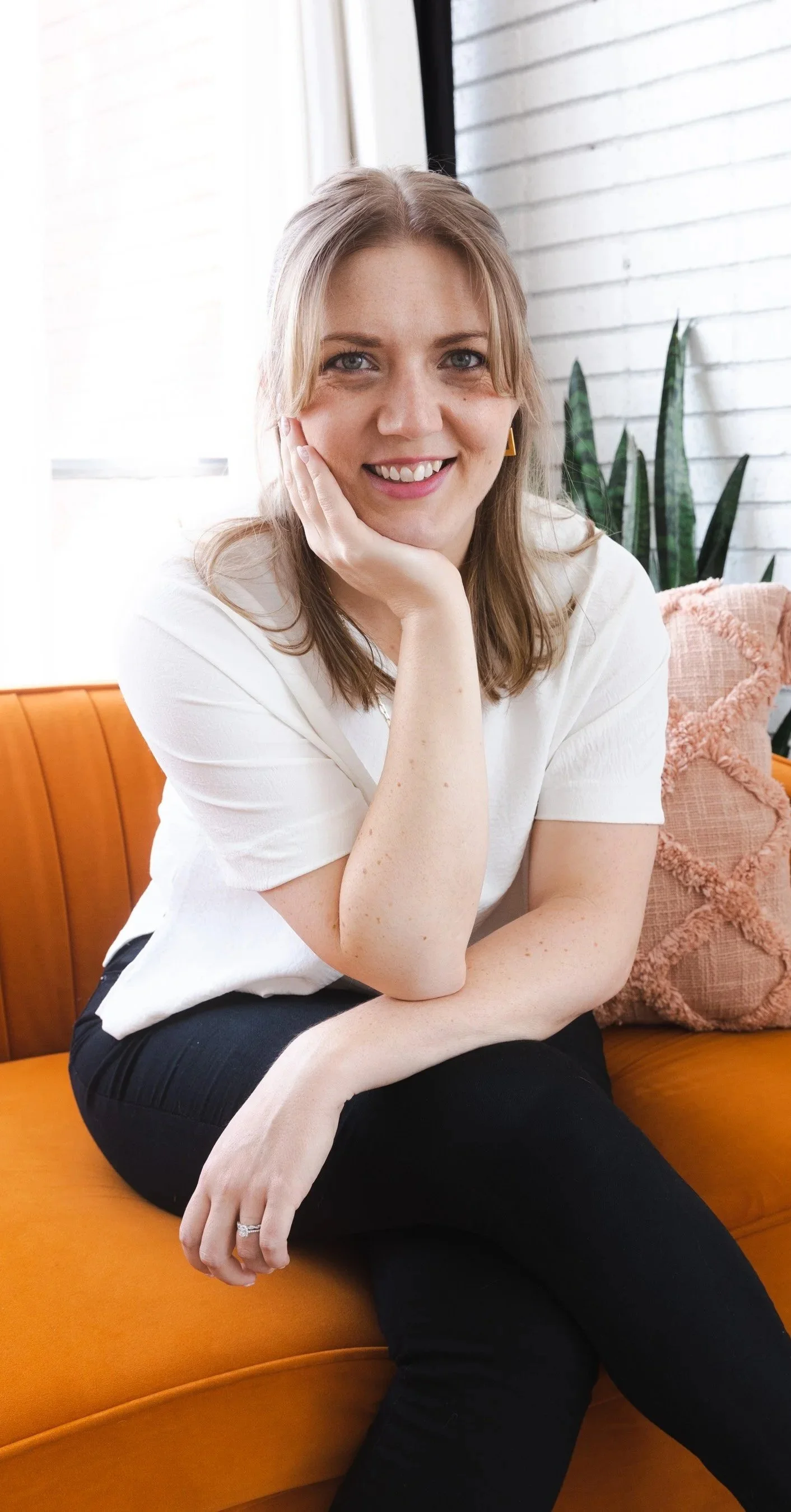 A woman with blonde hair, wearing a white top and black pants, sitting on an orange couch with a pink cushion, in front of a white brick wall with a plant in the background.