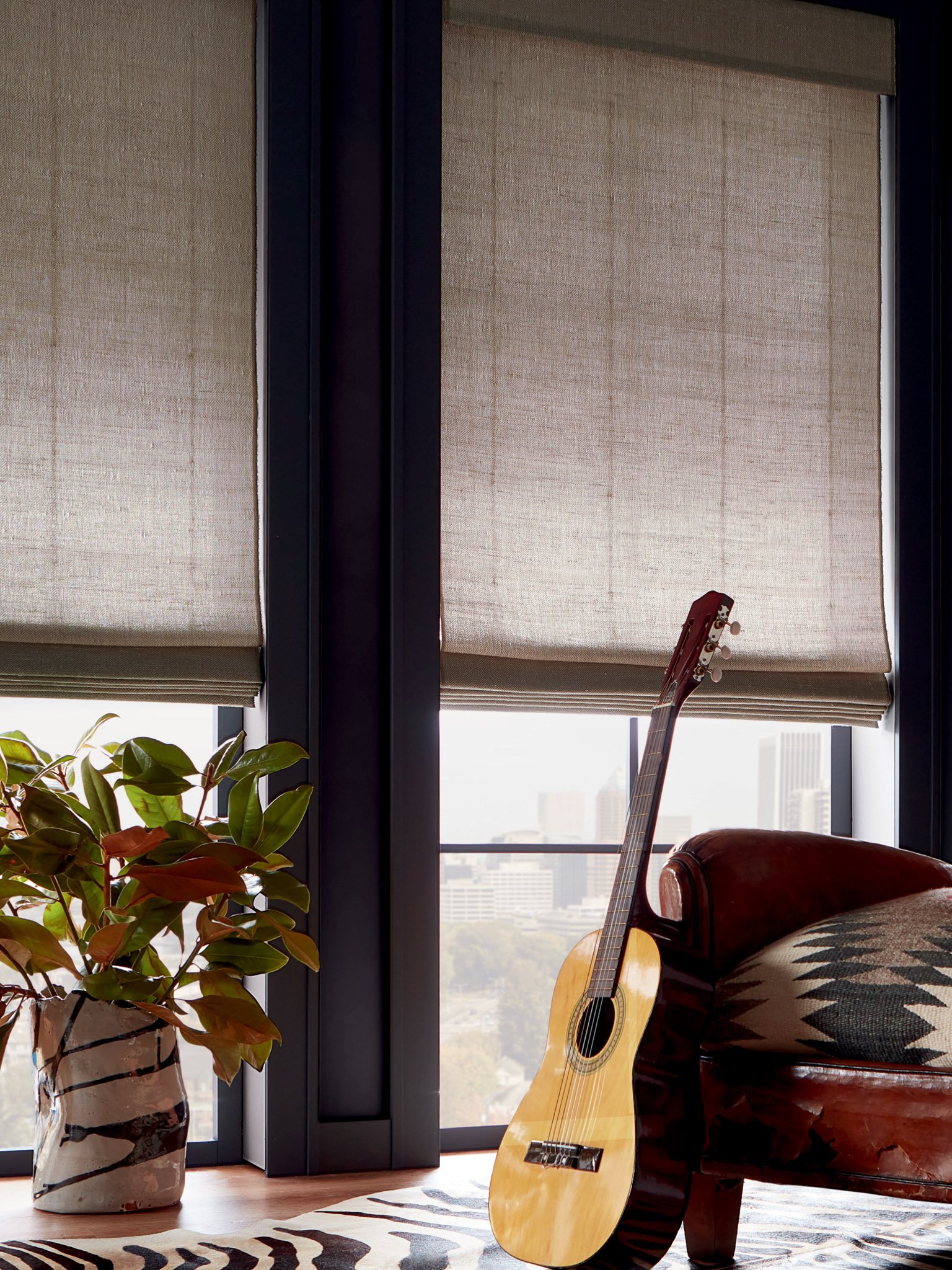 Room with floor-to-ceiling windows, a potted plant, an acoustic guitar leaning against a leather chair, and light-colored shades.