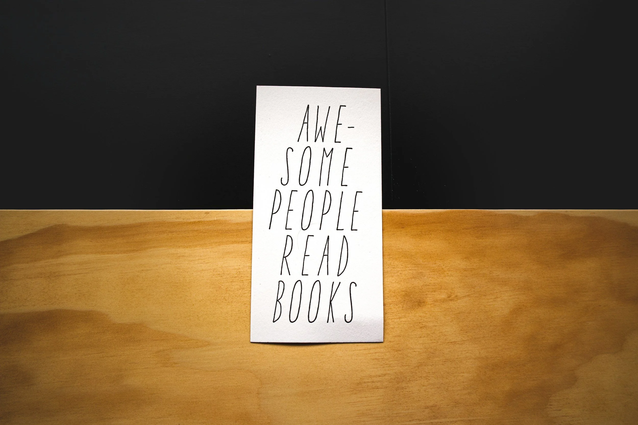 Card with "Awesome People Read Books" on a wooden surface with black background.