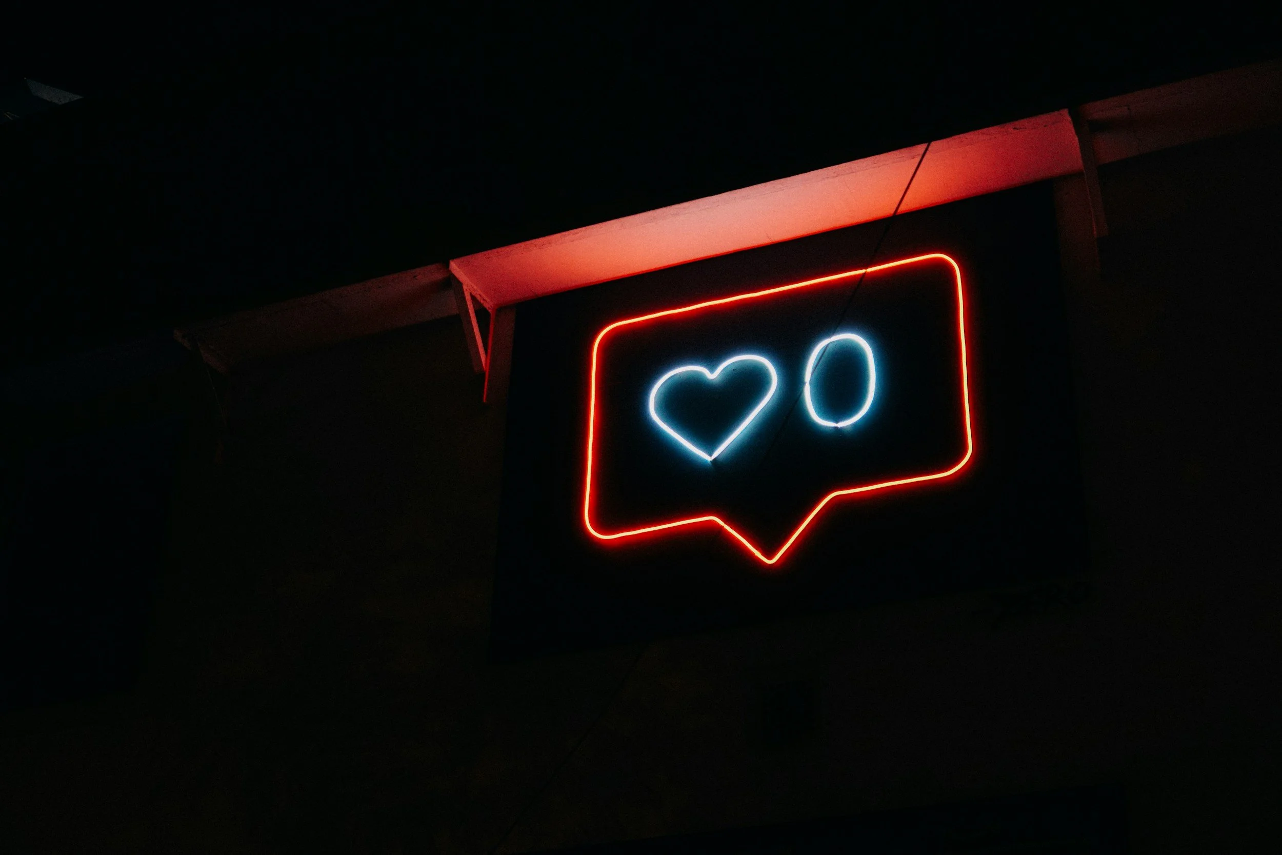 Neon sign resembling a social media notification with a red border, blue heart, and blue number zero inside a dark background.