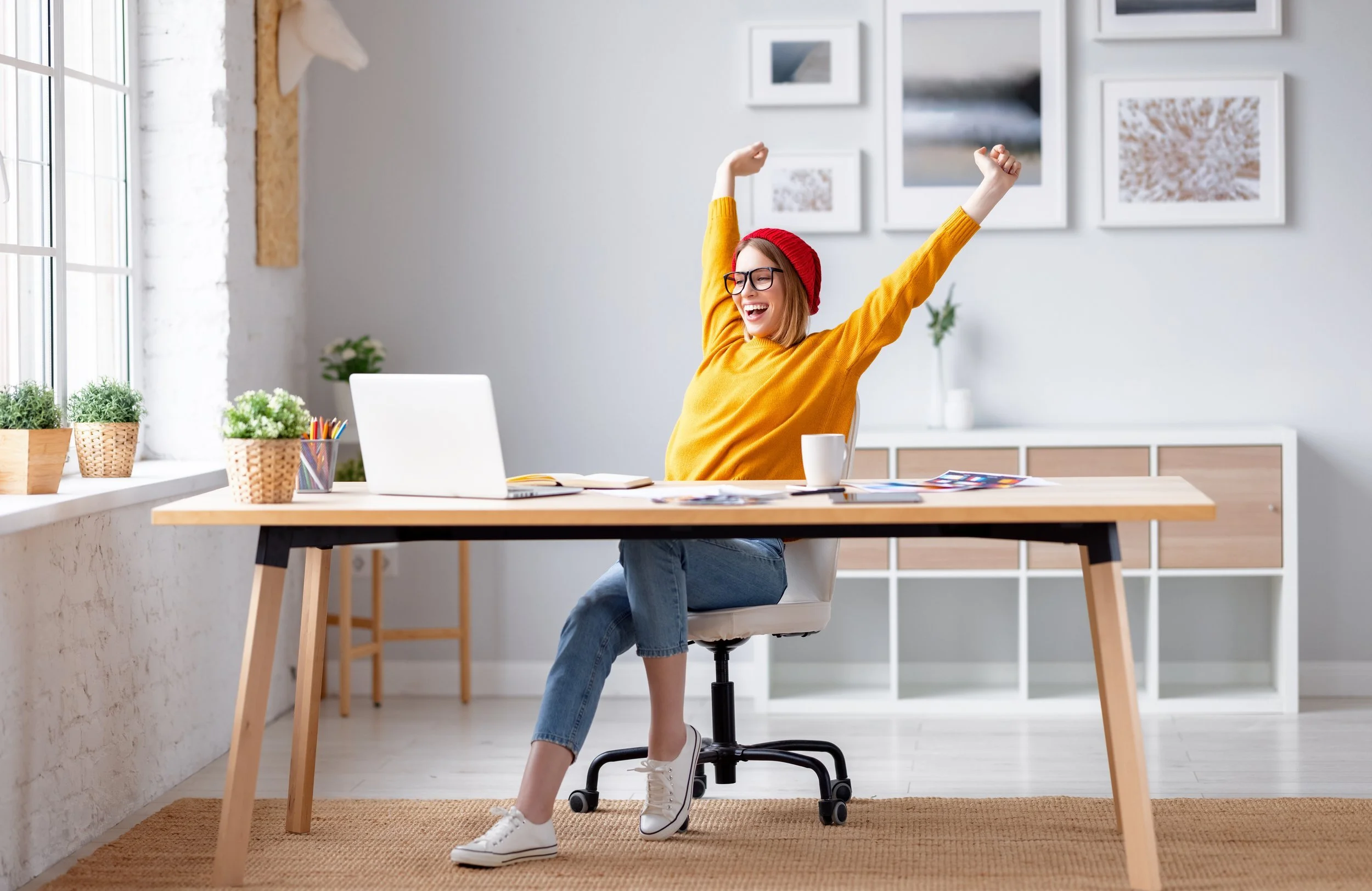 Woman in a yellow sweater and red beanie sitting at a desk with her arms raised in celebration, smiling in a bright, modern home office.