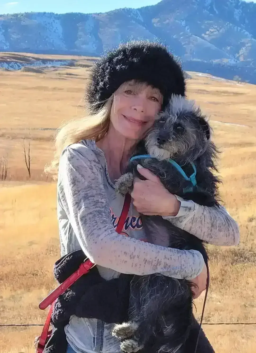 An image of owner, acupuncturist and herbalist Jen Katz in a winter hat holding a scruffy black and gray dog in an open field with mountains in the background.