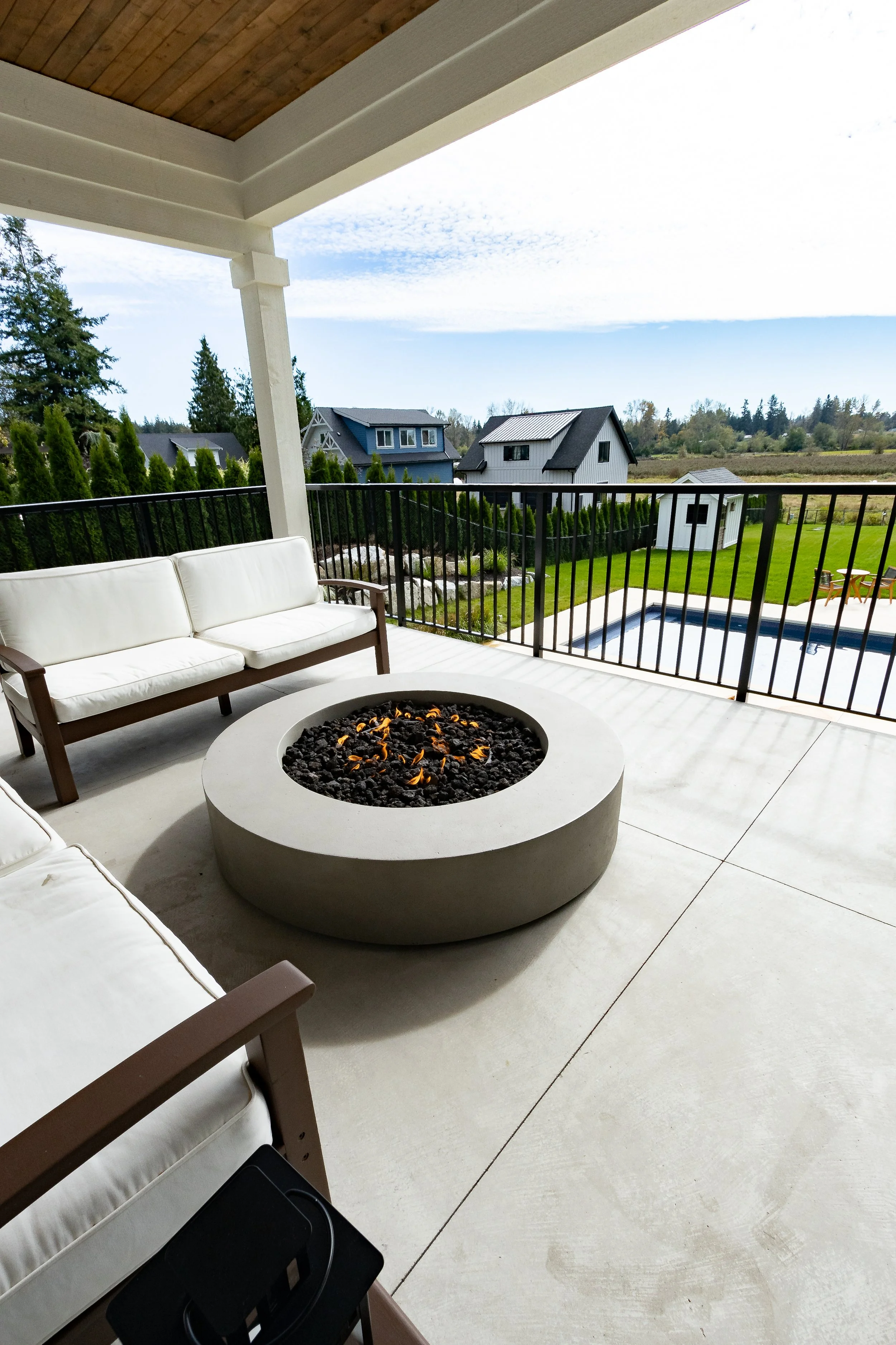 Covered outdoor patio with white cushioned seating, a modern fire pit with black stones and flames, and a view of houses, green lawn, and a swimming pool beyond the fence.