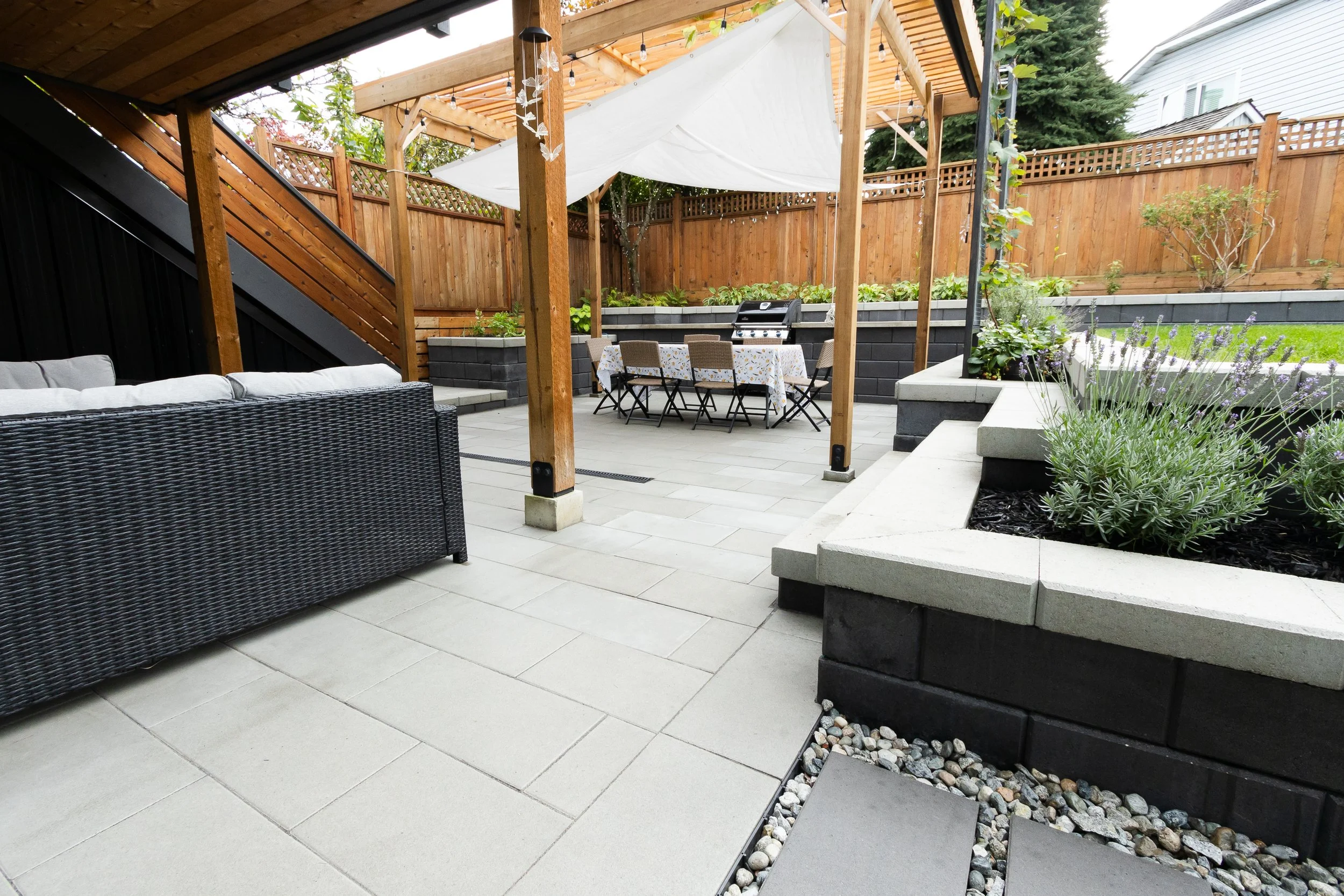 A backyard patio with outdoor furniture, including a black wicker sofa with white cushions, a dining table with chairs and a tablecloth, and a grill in the background. There are flower beds with plants and a wooden fence surrounding the yard.