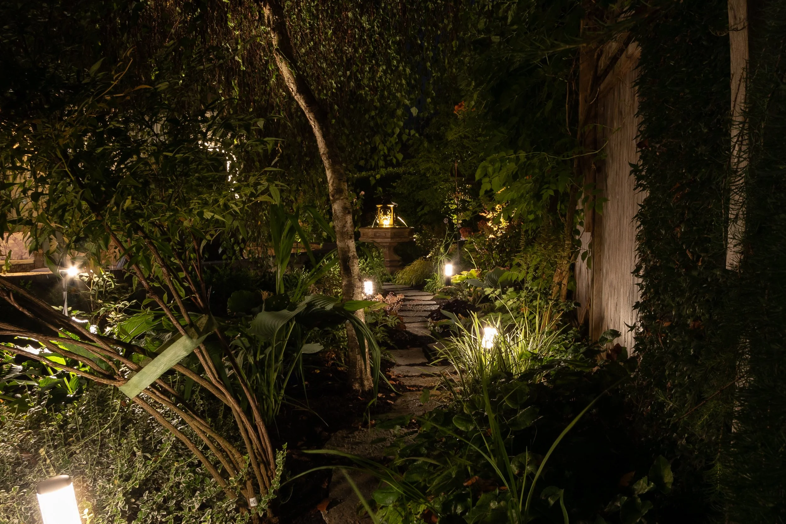 A nighttime garden scene illuminated by pathway lights, featuring lush greenery, trees, and a stone pathway leading to a decorative fountain.