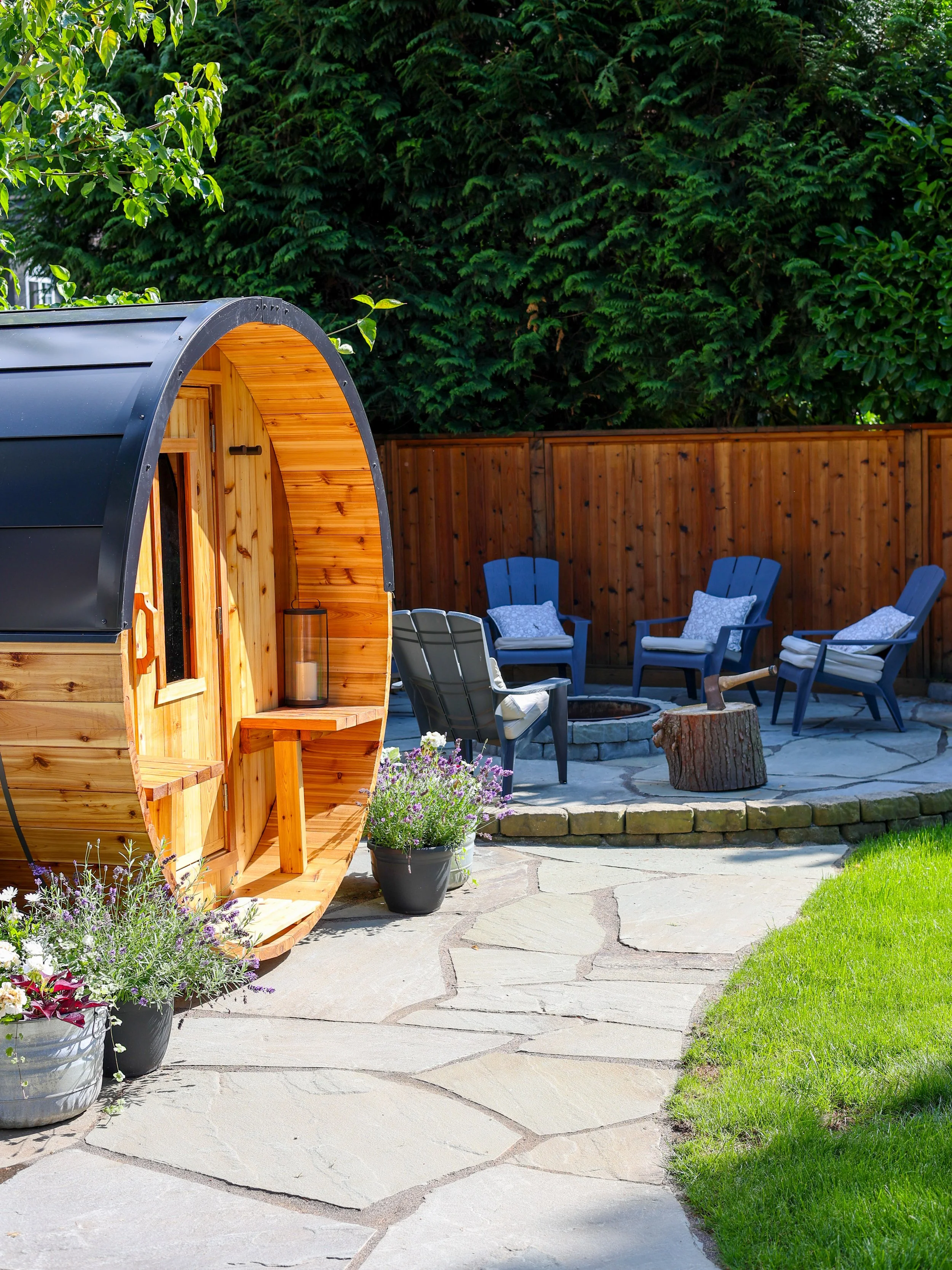 A backyard patio with a wooden sauna, surrounded by potted flowers, a fire pit, and four blue chairs with cushions on a stone-paved area, with trees and a wooden fence in the background.