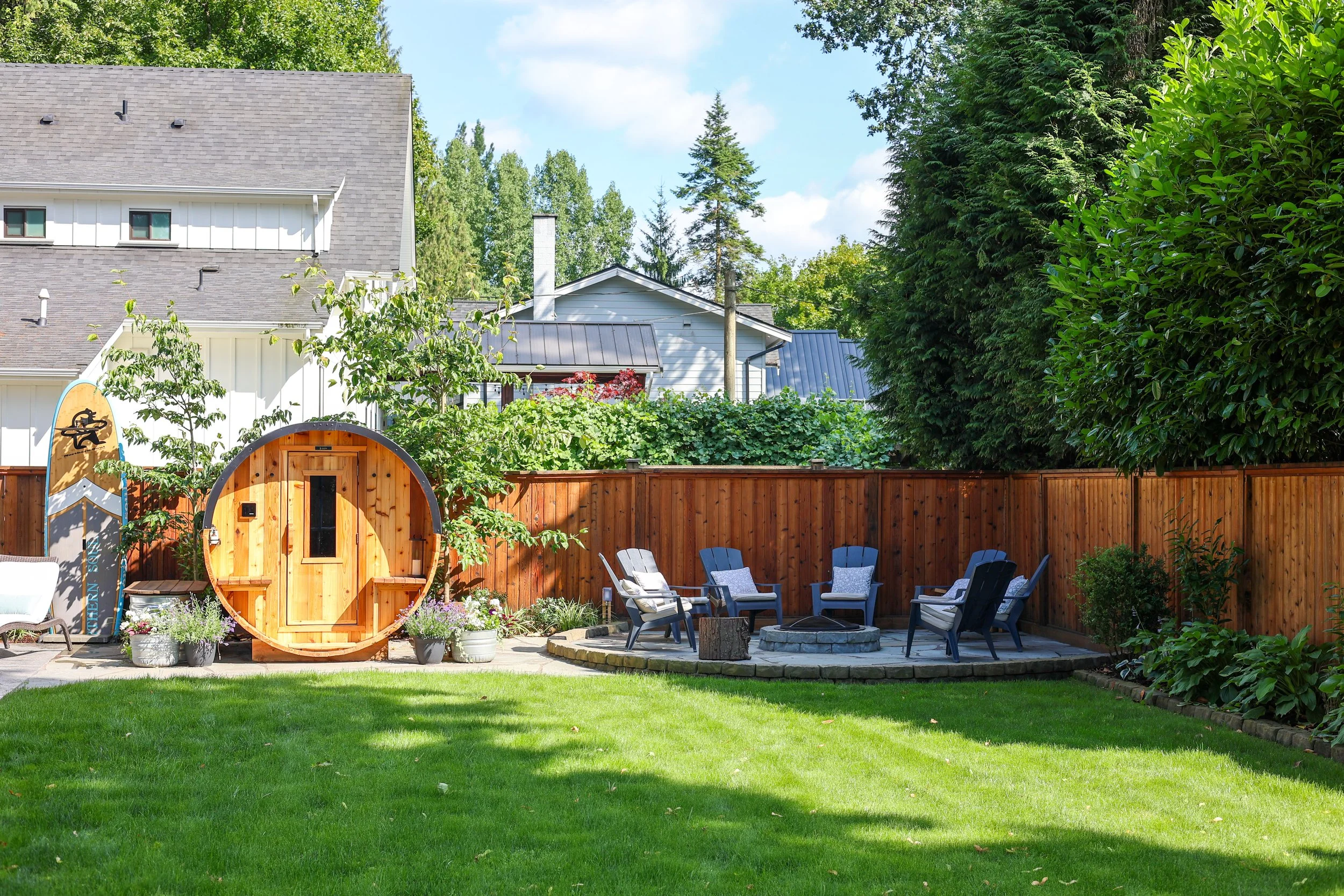 Backyard with chain-link fence, six blue and white chairs arranged around fire pit, small wooden sauna, lush green grass, and various trees and plants.