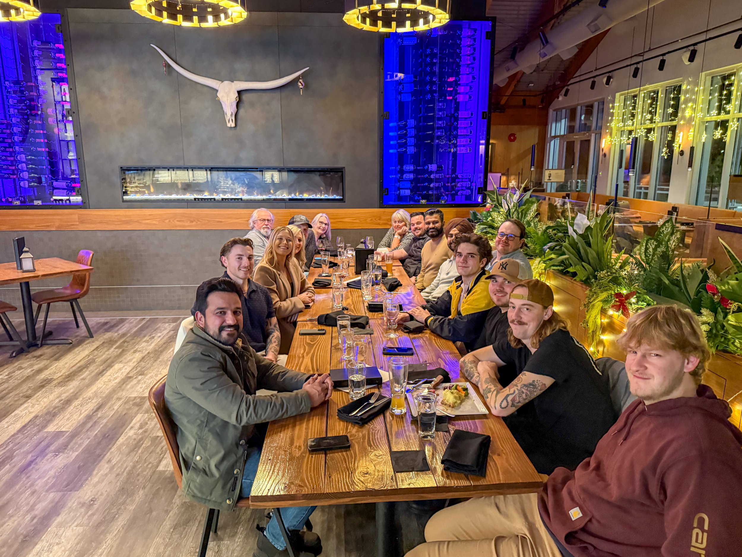 Group of people sitting at a long wooden table in a restaurant, with festive holiday decorations and large green plants behind them, and a modern interior with a fireplace and decorative lighting.