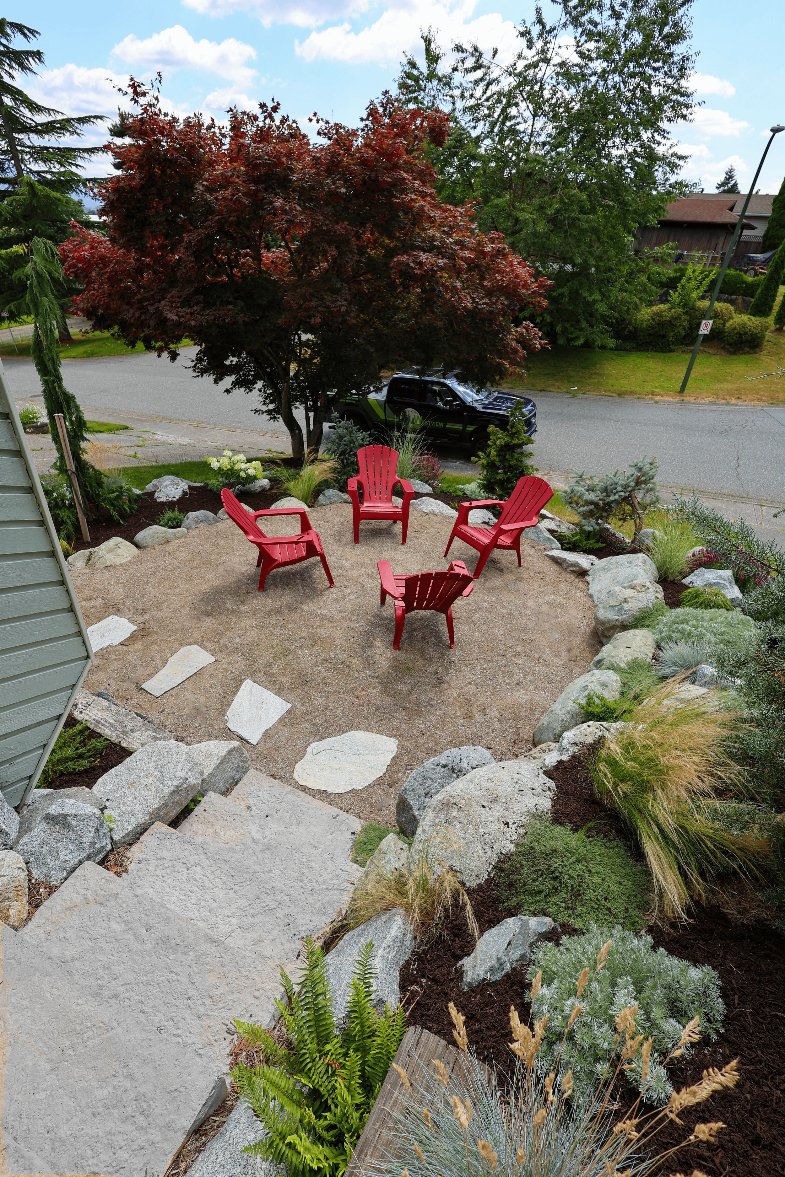 Backyard garden with four red Adirondack chairs arranged in a circle on a small dirt area, surrounded by rocks and various plants, with a large red-leafed tree and a street with a black vehicle in the background.