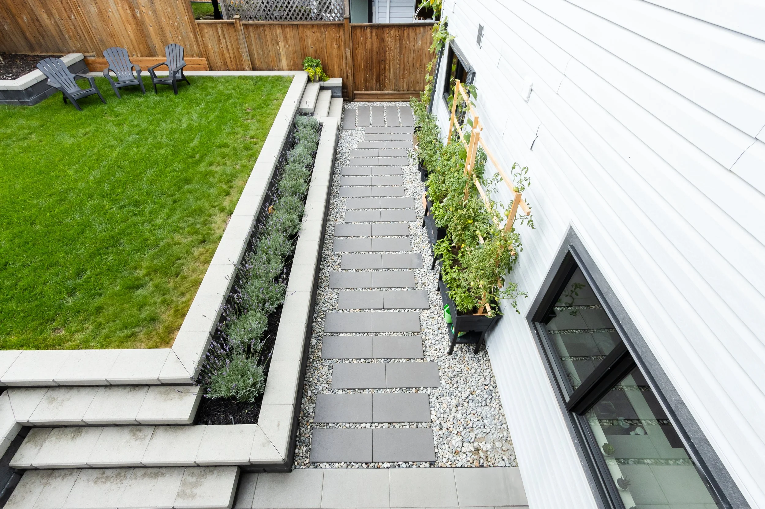 A backyard with a grassy lawn, a wooden fence, outdoor seating, and a garden area with potted plants and a stone pathway.