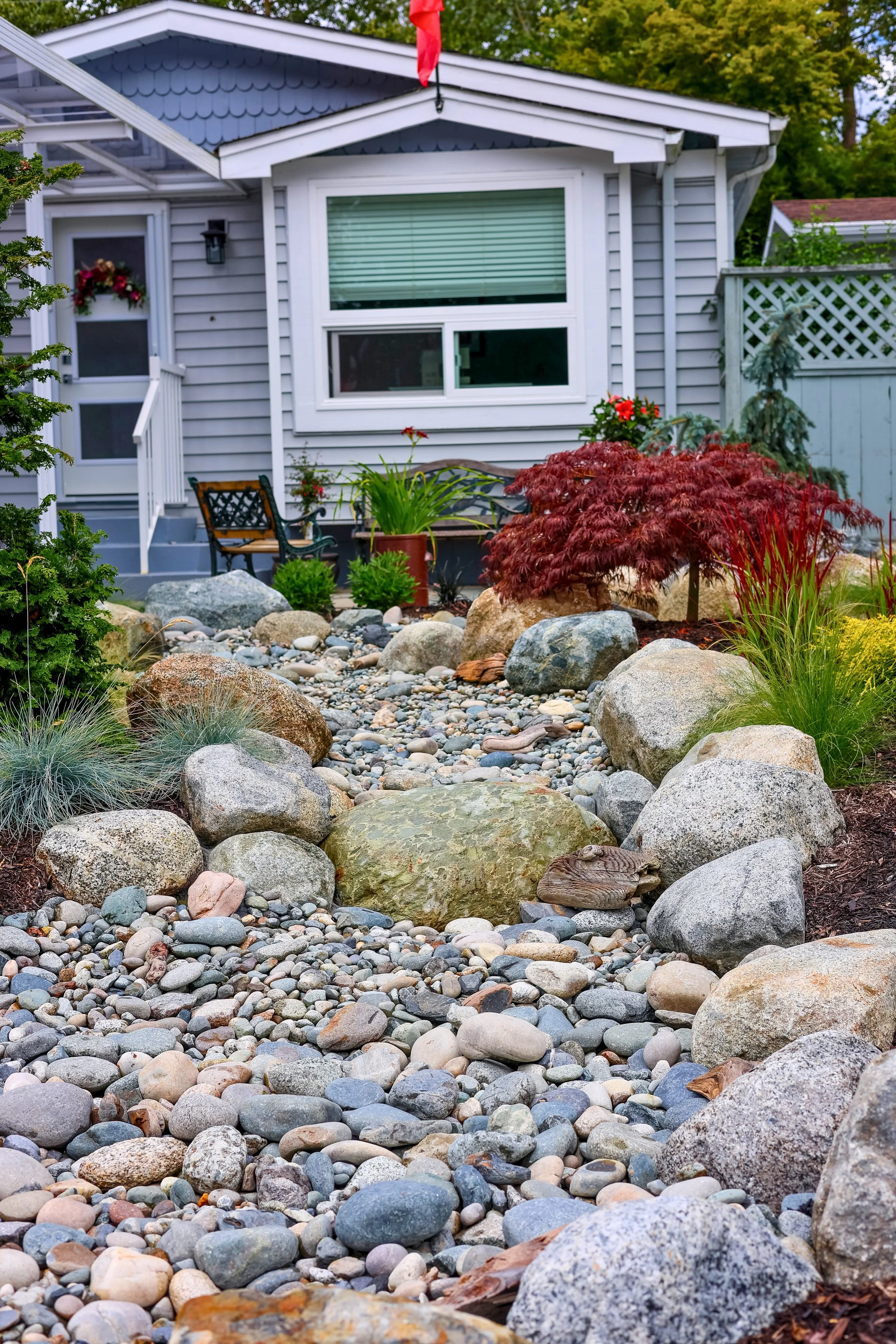 Rock garden with large rocks, pebbles, and plants in front of a gray house with large window, porch, and black chairs.
