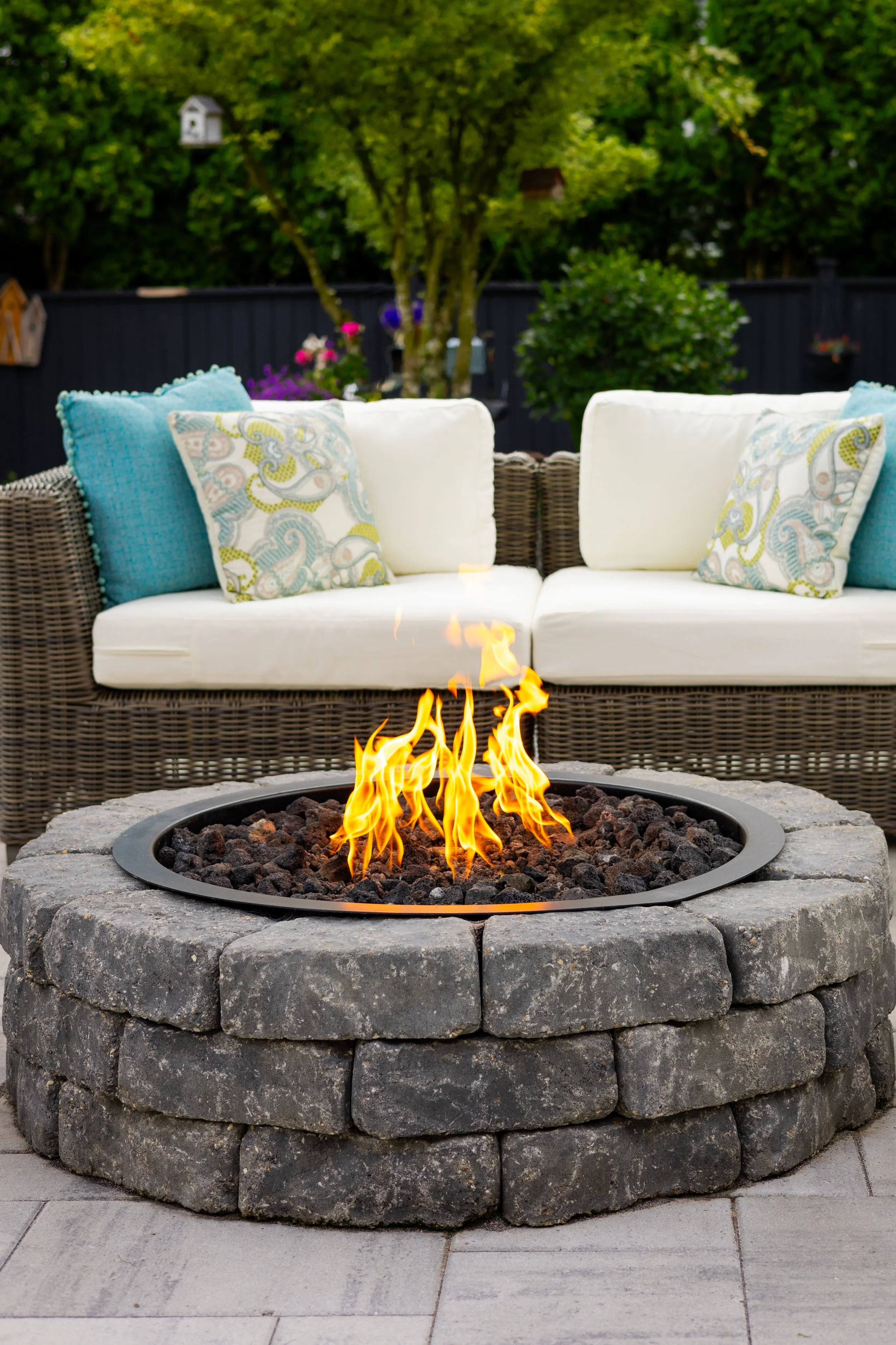 Outdoor patio with a circular stone fire pit and a wicker sofa with white cushions and colorful pillows, surrounded by greenery.