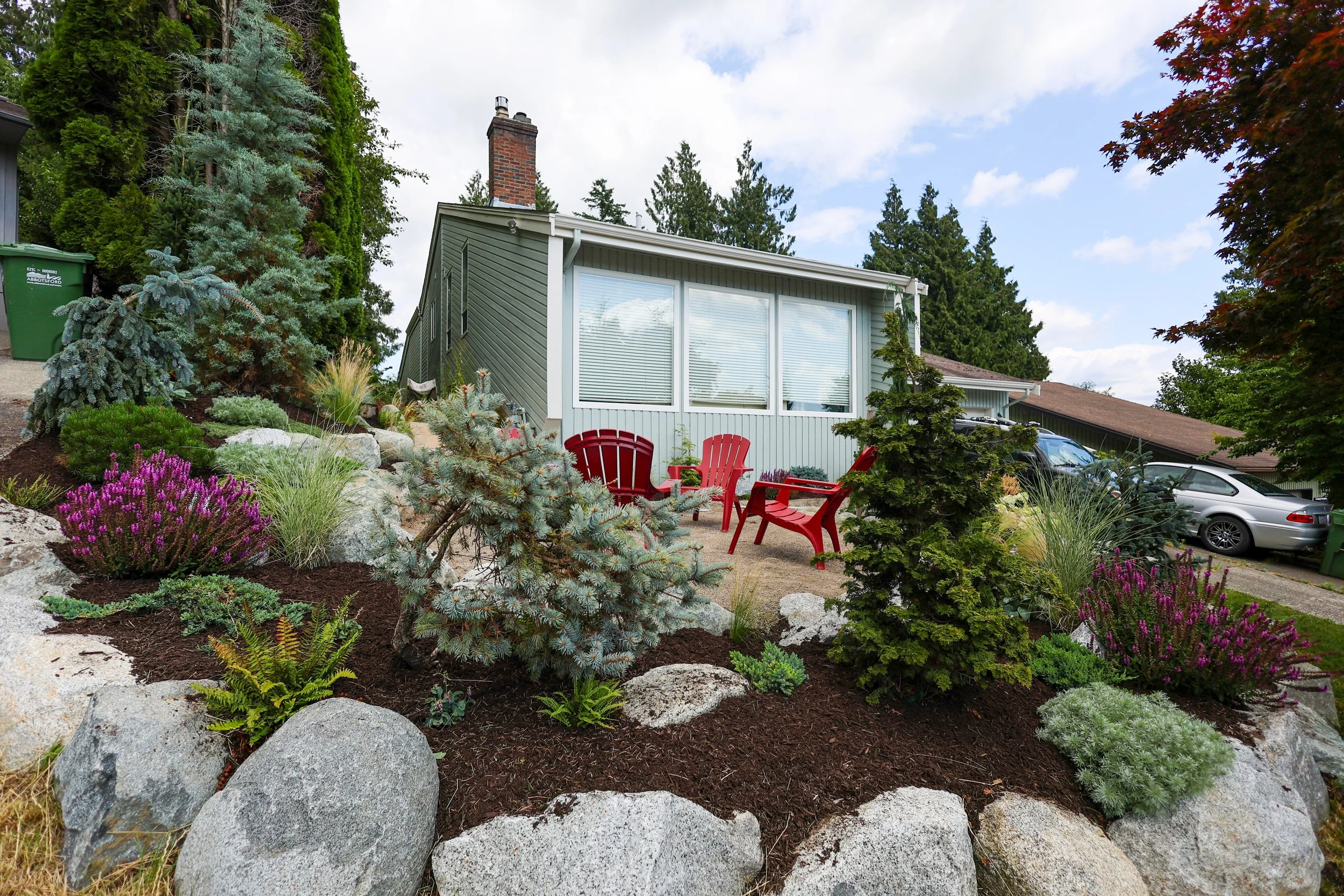 A house with a small garden area featuring colorful plants and shrubs, including pink and purple flowers, a small pine tree, and large rocks, with red outdoor chairs and a car parked nearby.