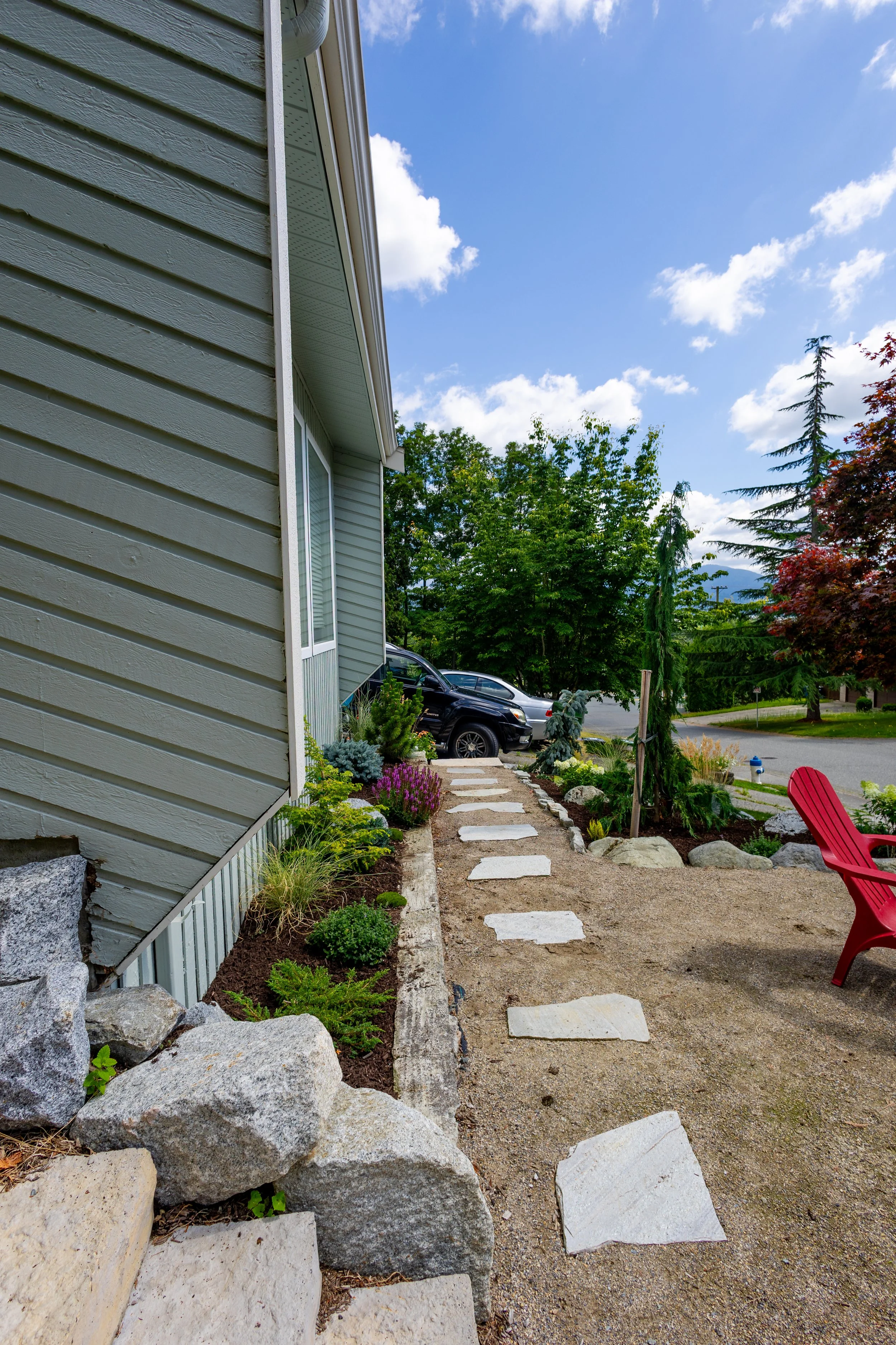 A landscaped front yard with a stone pathway beside a house, featuring plants, rocks, and a red chair, with a street, parked cars, and trees under a partly cloudy sky.