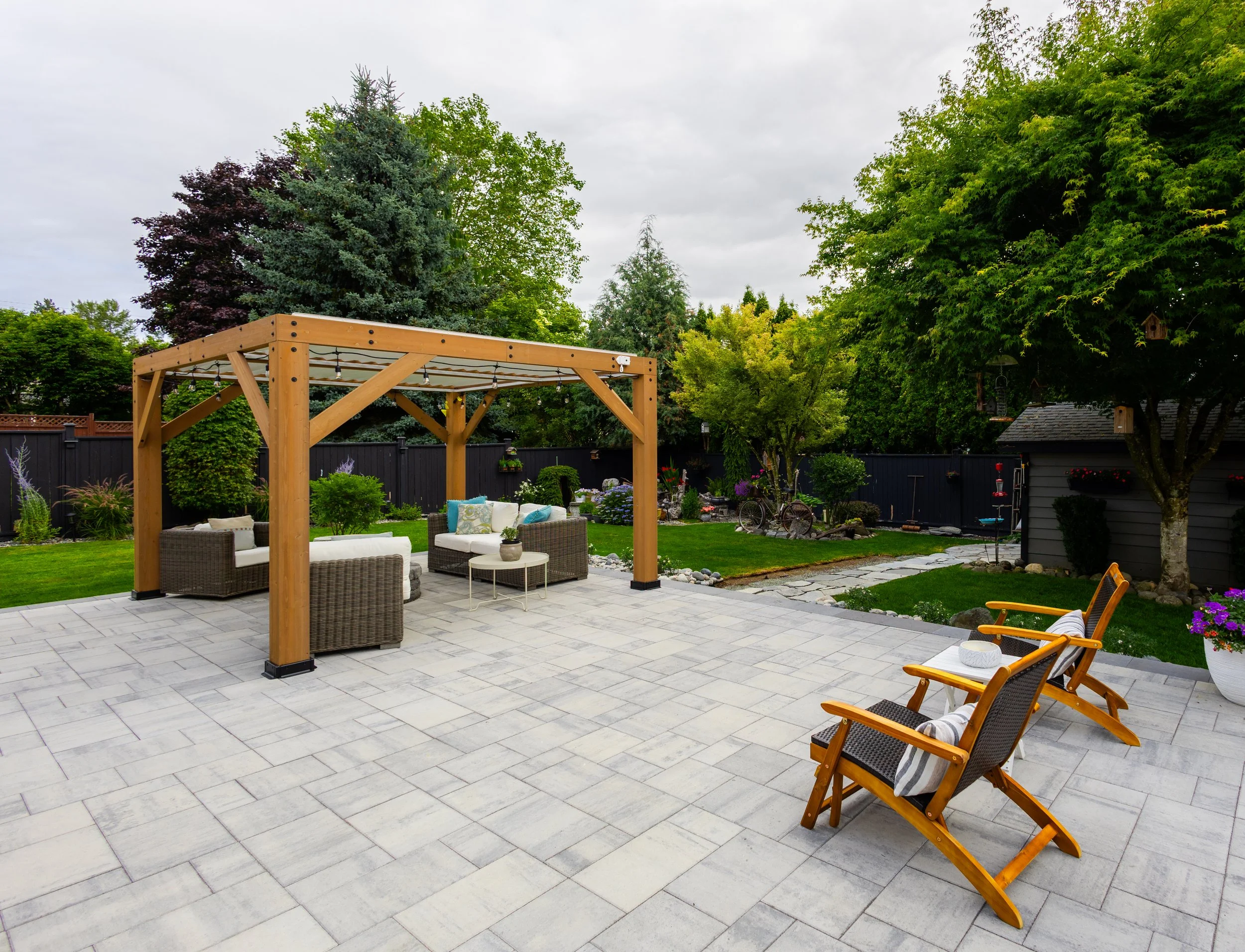 A spacious backyard patio with outdoor furniture under a wooden pergola, surrounded by lush green trees and landscaped garden.