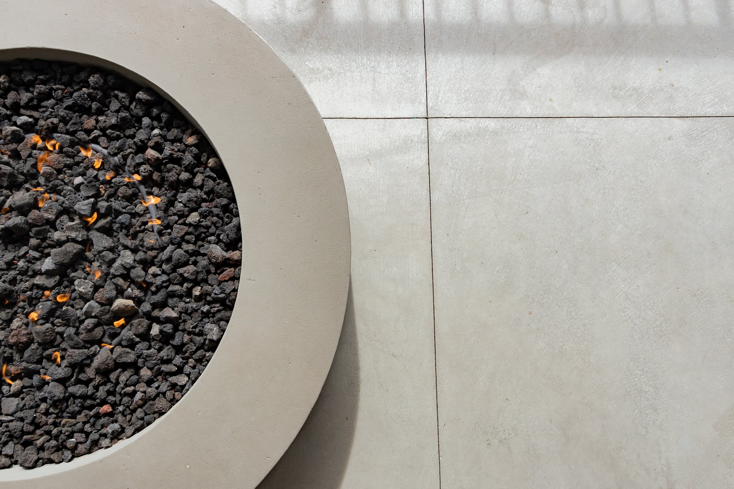 Top view of a circular concrete planter filled with black volcanic rocks and small orange flowers, placed on a light-colored tiled floor.