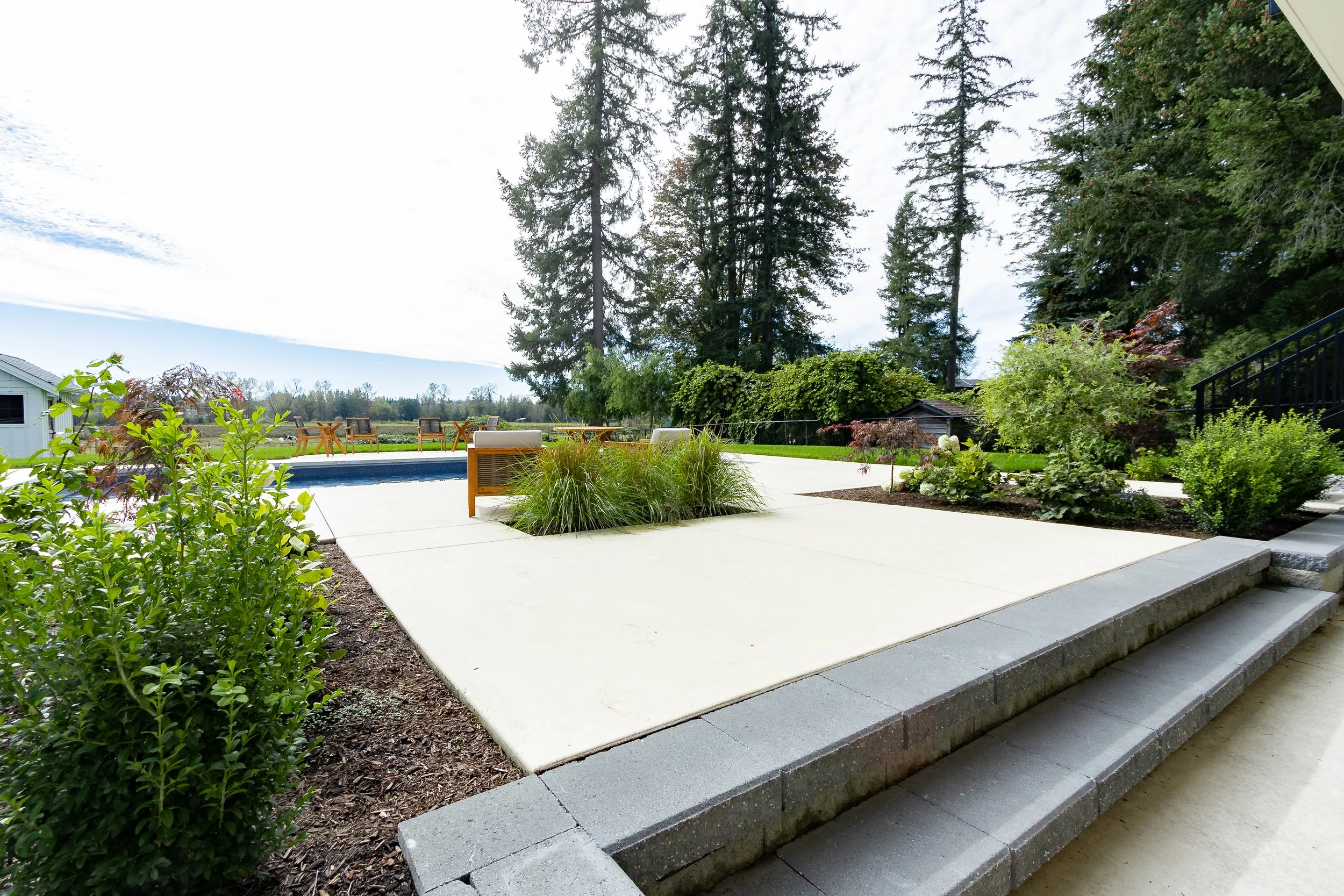 A backyard patio with concrete paving, garden beds, and outdoor furniture, surrounded by trees and greenery.