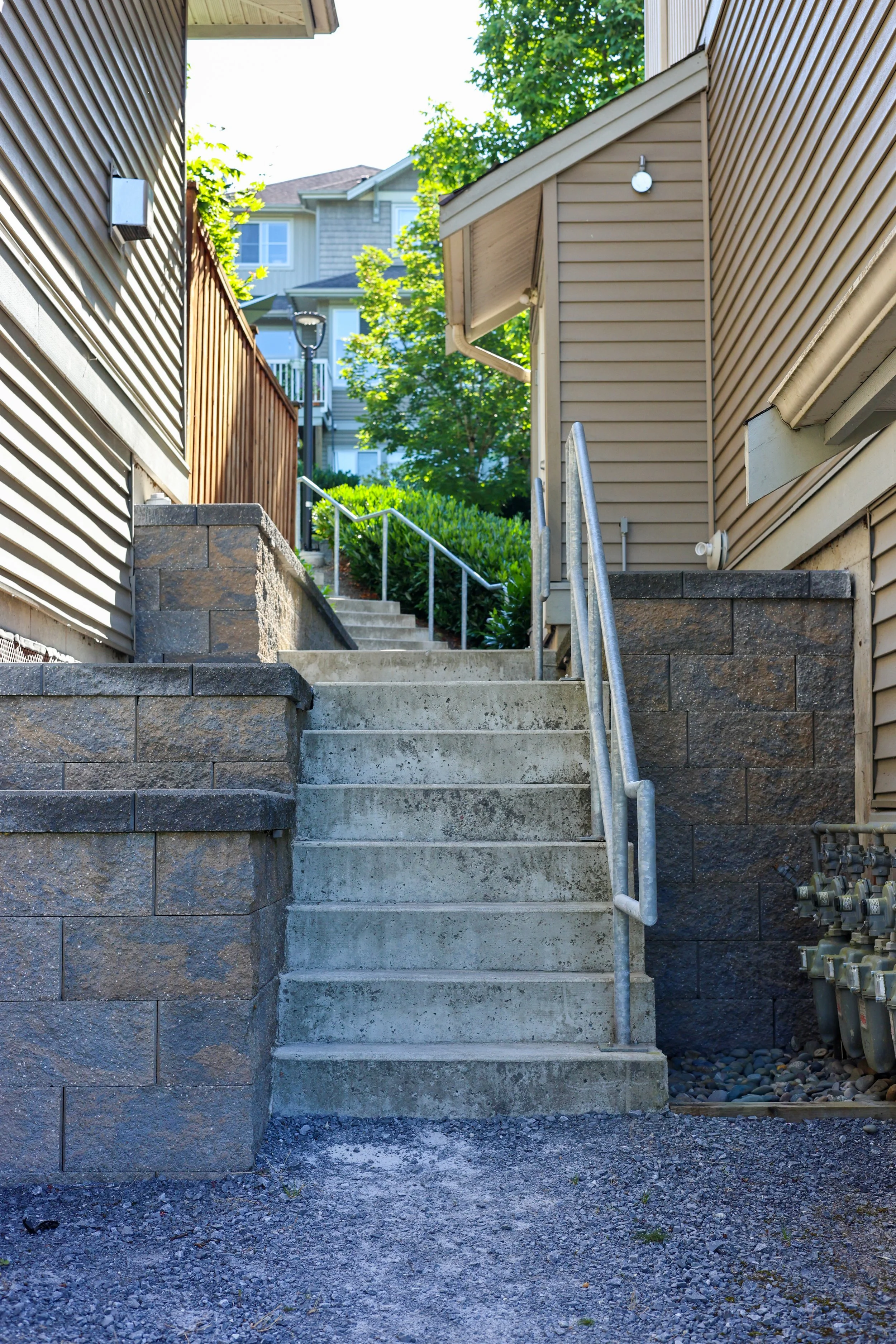 Concrete stairs with metal handrails leading up between two houses in a residential area, with greenery and trees in the background.