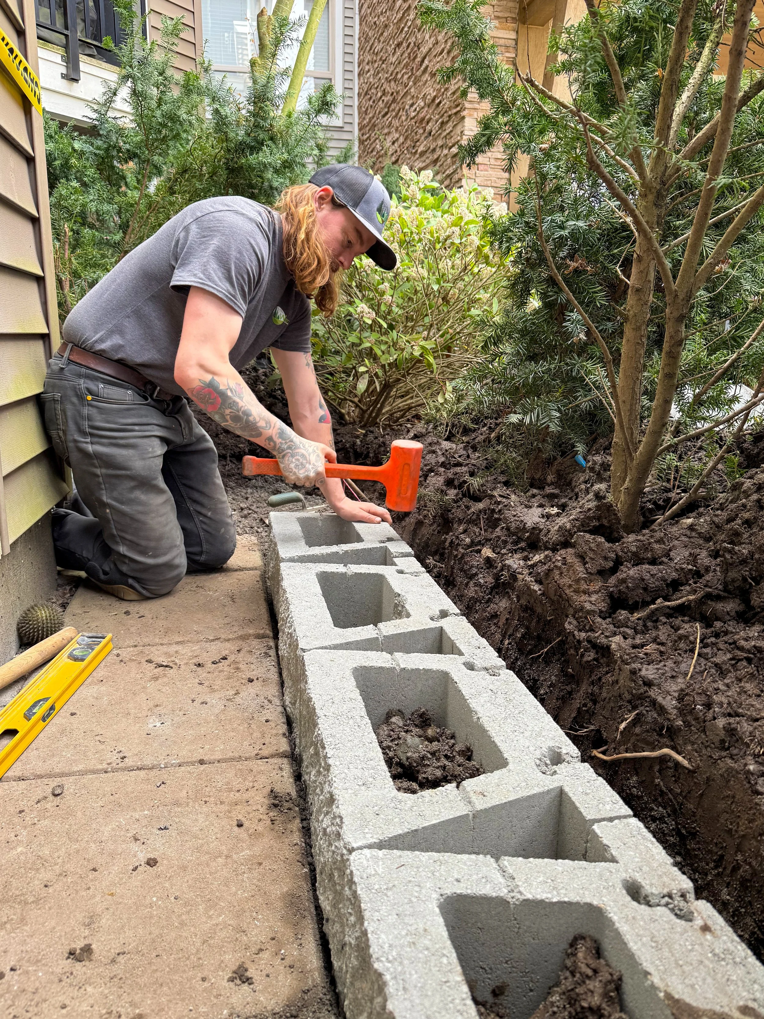 A man kneeling on a concrete patio, using a hammer to build a cinder block wall beside a garden with trees and bushes.