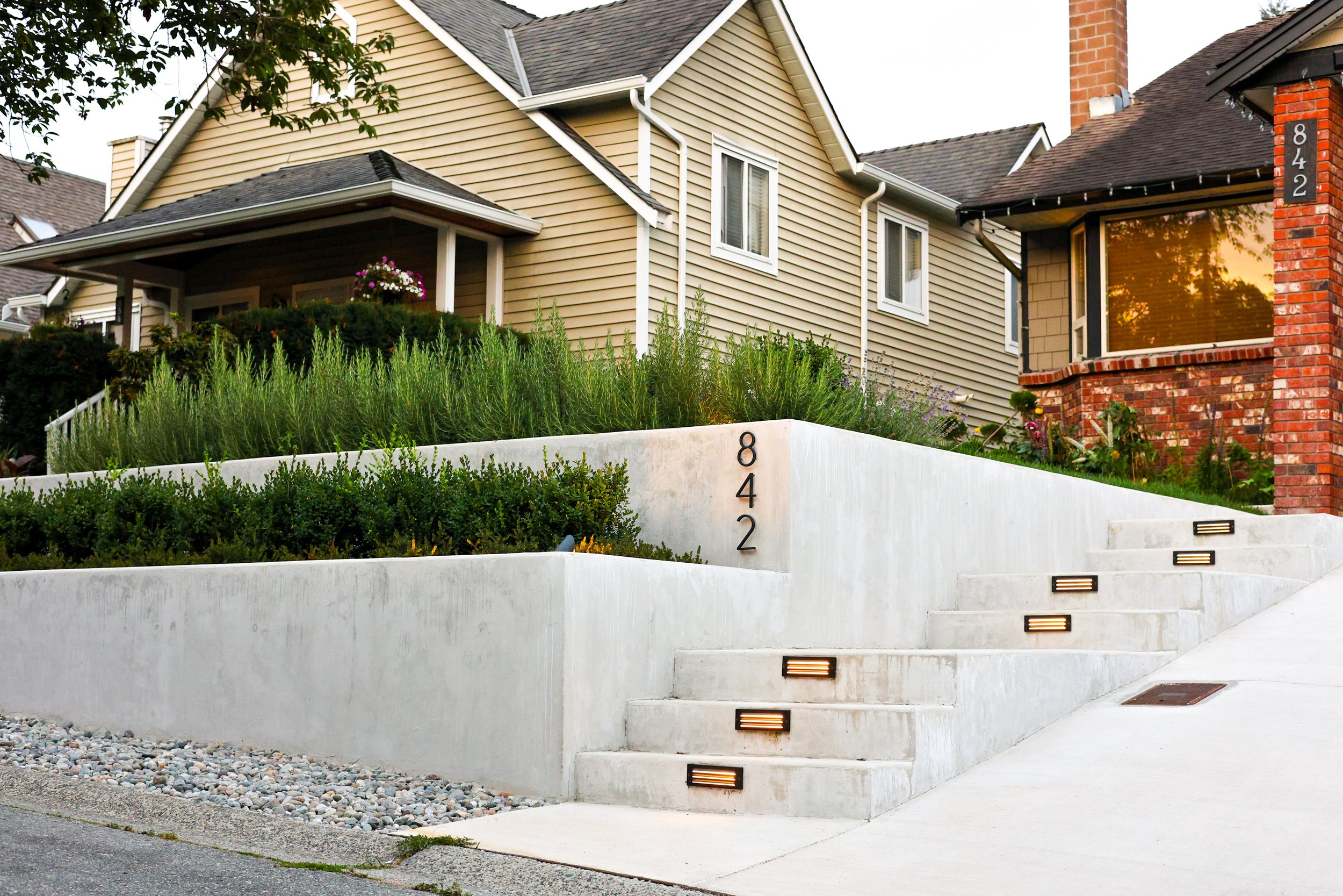 Front view of a residential house with a concrete staircase leading up to the front door, displaying the house number 842 on the wall and steps with integrated lighting, surrounded by landscaped greenery and a driveway.