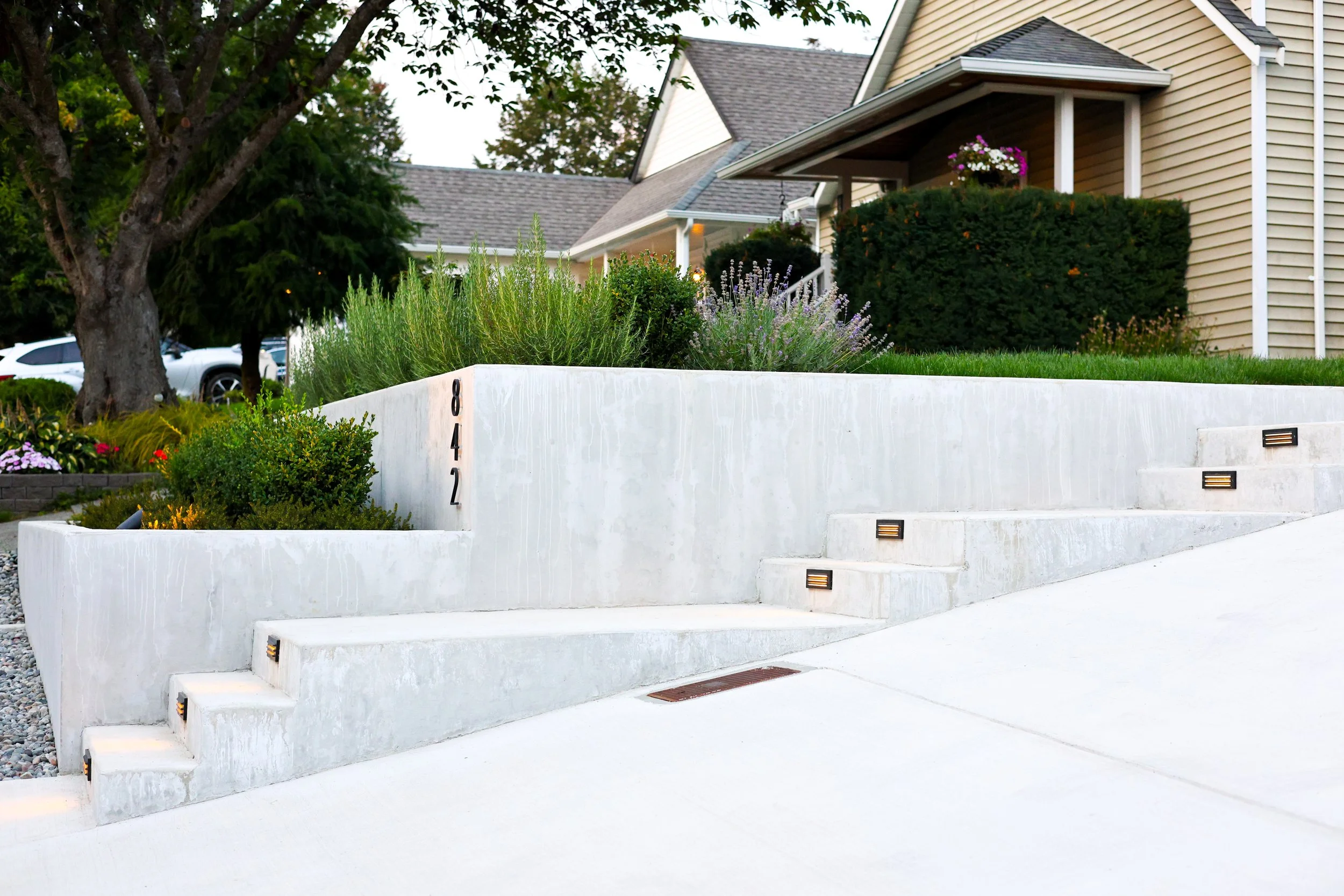Modern residential front yard with concrete stairs, landscape lighting, and house with beige siding and a porch with hanging flower basket.