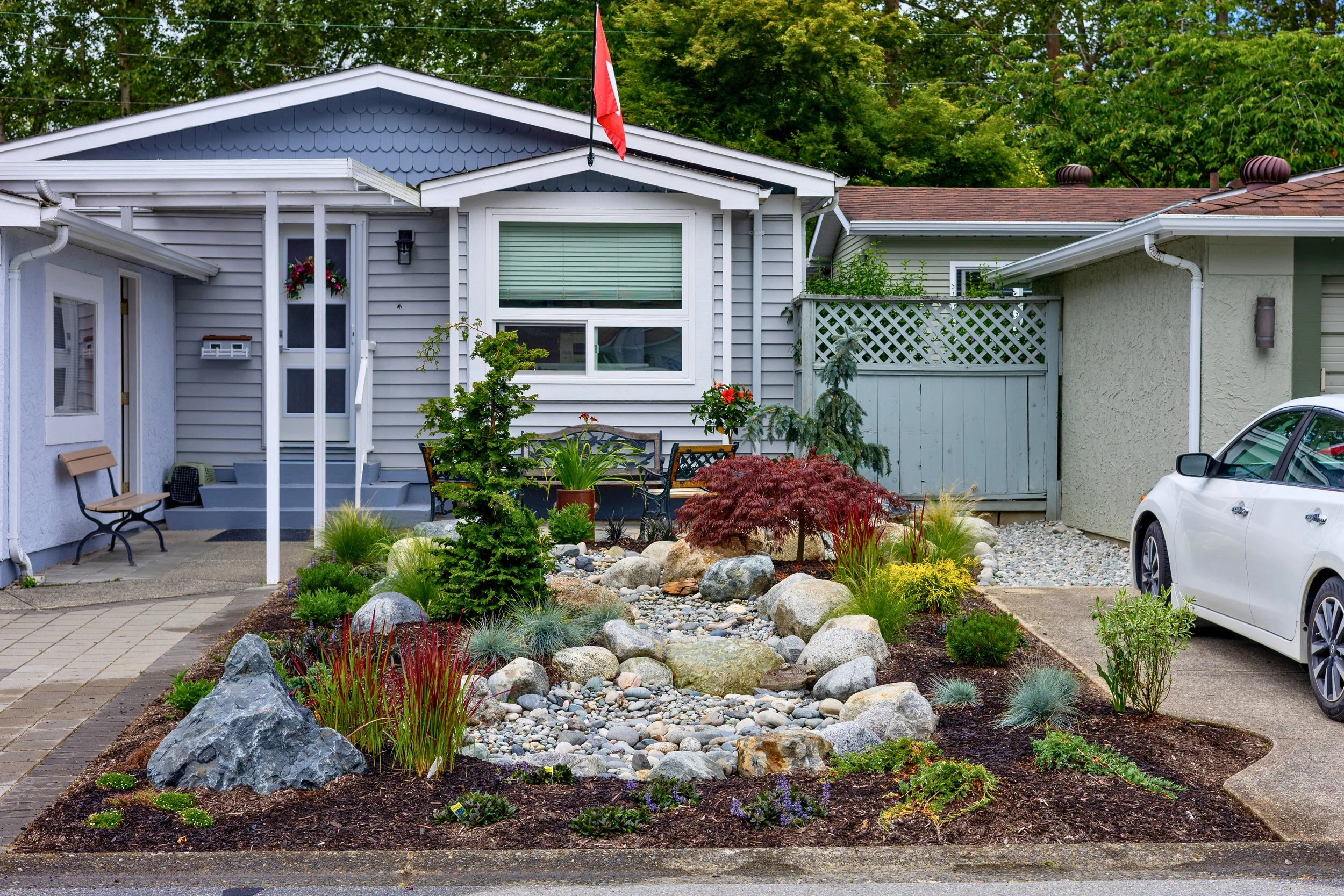 Front yard with rock garden, plants, trees, and a house with a porch and a parked white car.
