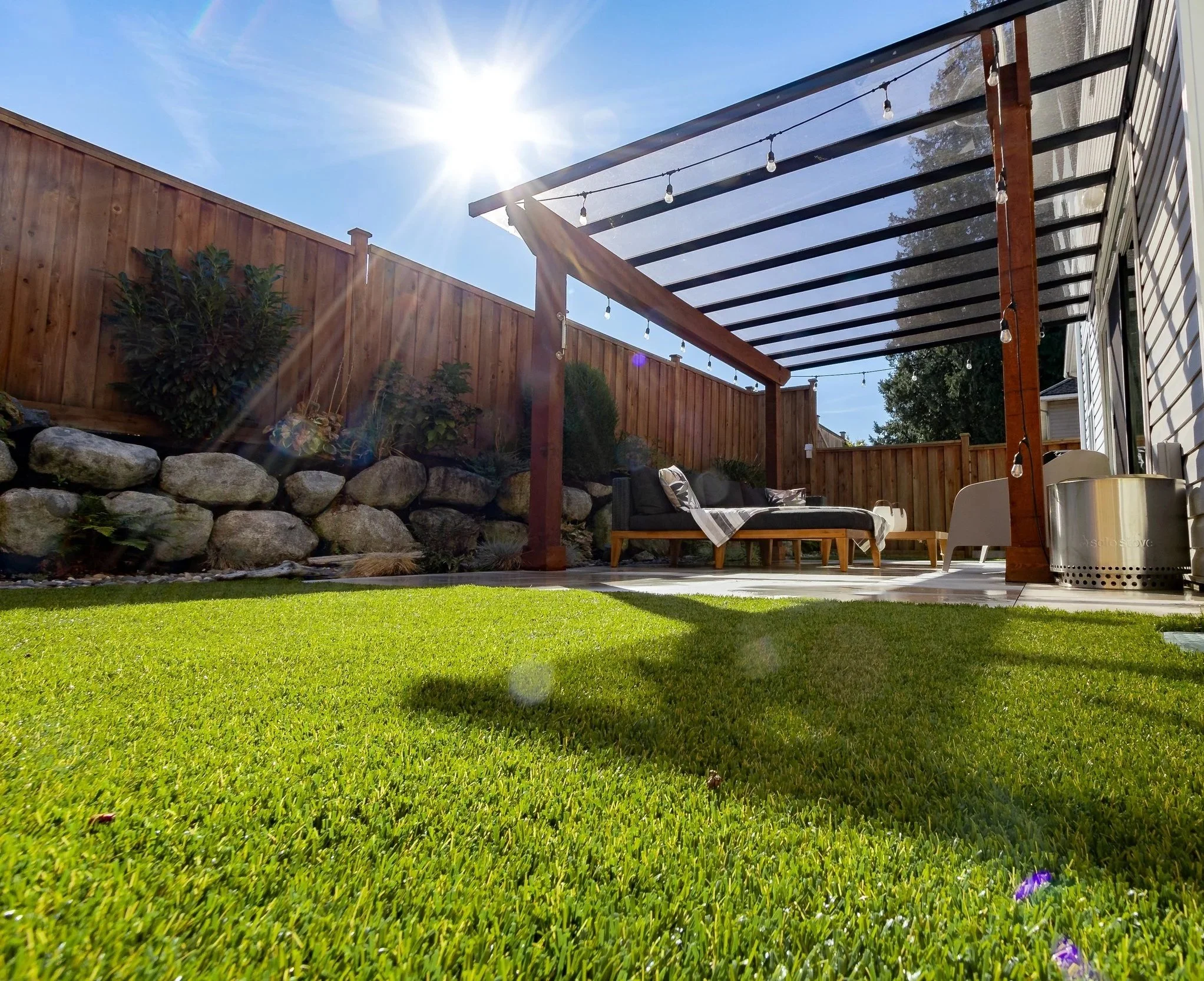 Backyard patio with green grass, wooden fence, rock garden, and a pergola with string lights. Outdoor seating includes a sofa and a chair, with bright sunlight and lens flare.