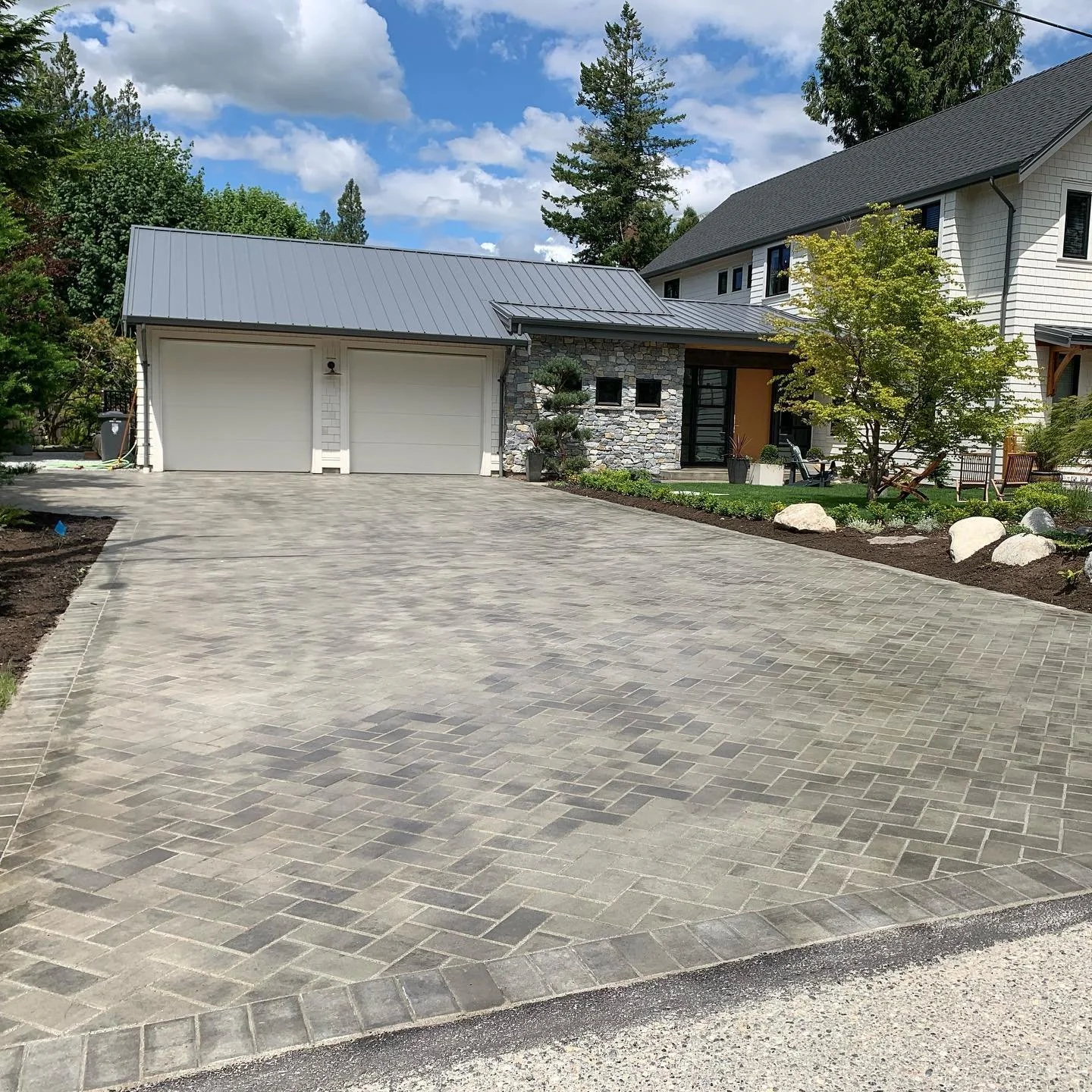 A modern house with a paved driveway, surrounding greenery, small trees, large rocks, and garden furniture under a partly cloudy sky.
