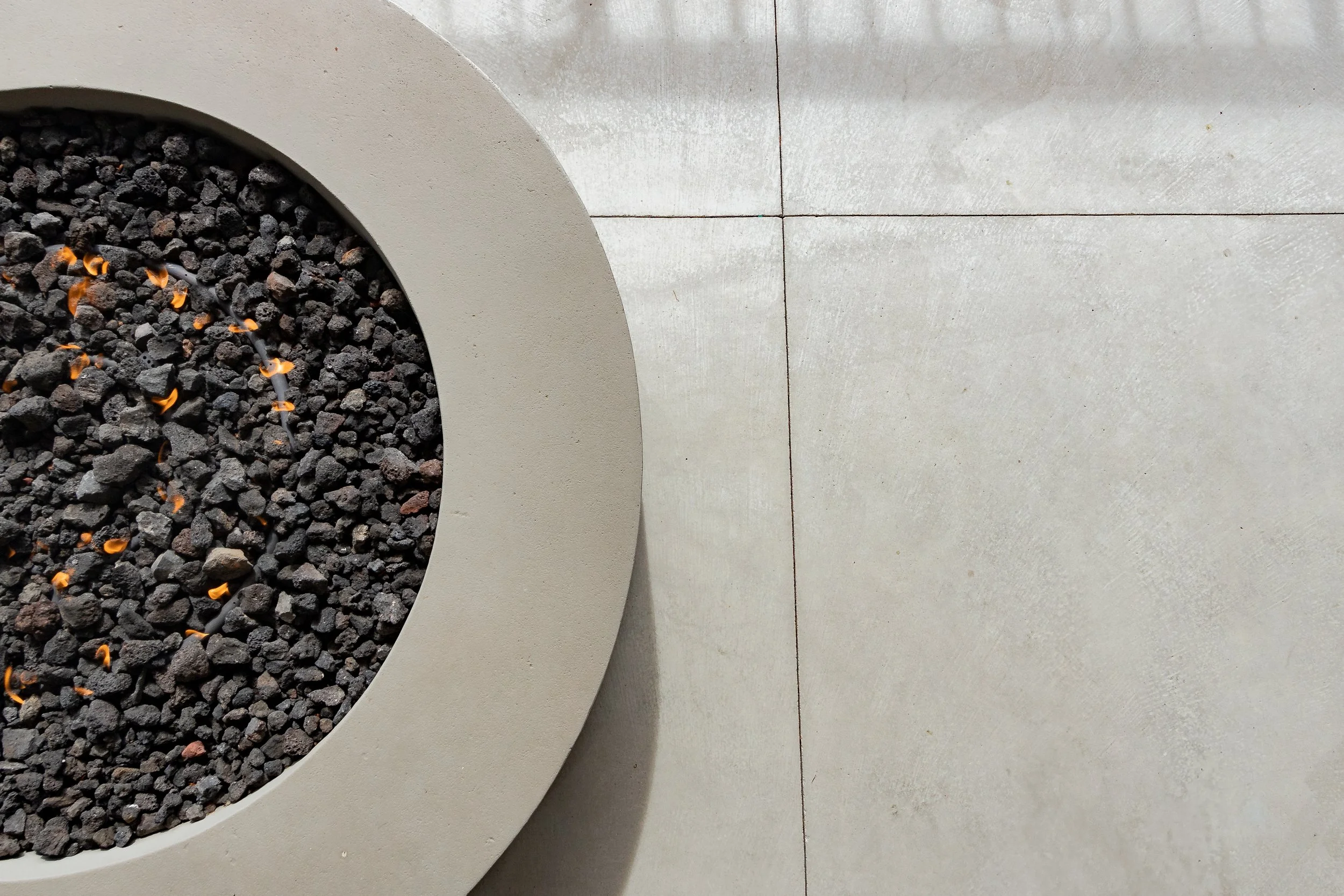 Top-down view of a circular planter with black rocks and small orange flower buds, adjacent to a tiled concrete floor.