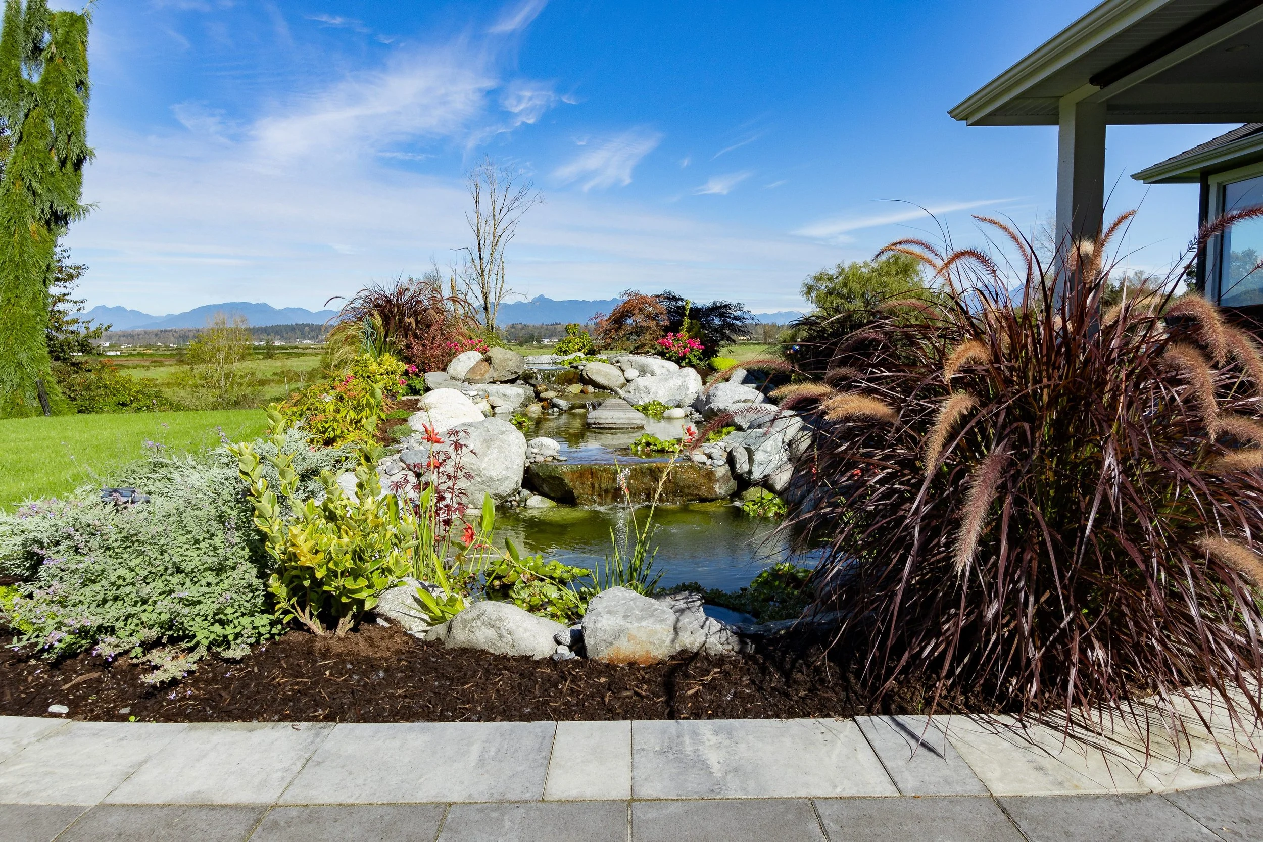 A landscaped garden with a small pond, rocks, and lush plants, overlooking mountains under a blue sky.