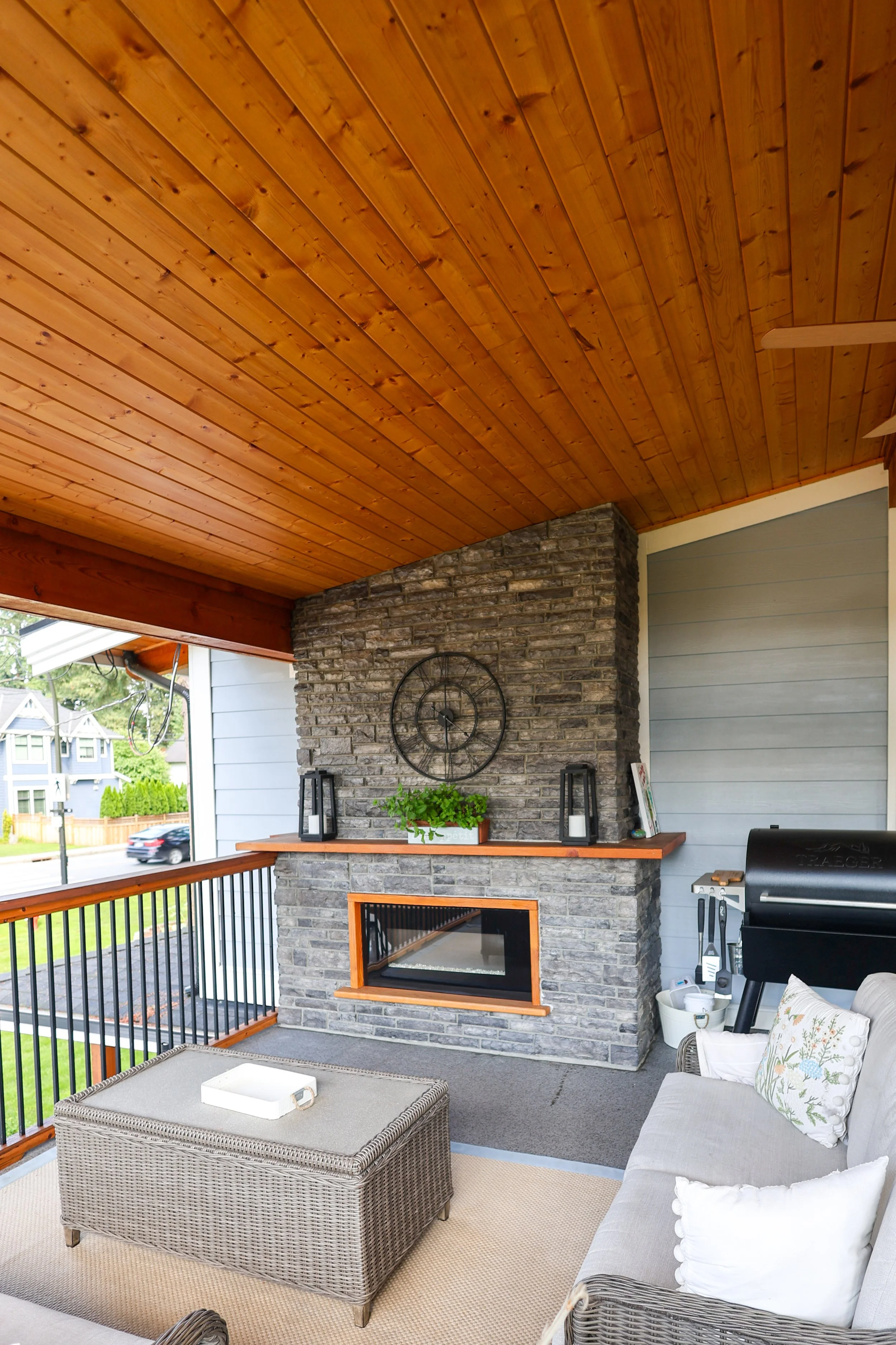 A cozy outdoor patio area featuring a brick fireplace with a mounted clock, a wood shelf decorated with lanterns, a potted plant, and other decor, a grill, outdoor furniture, a wicker ottoman, and a ceiling fan with wooden blades, with a view of neig