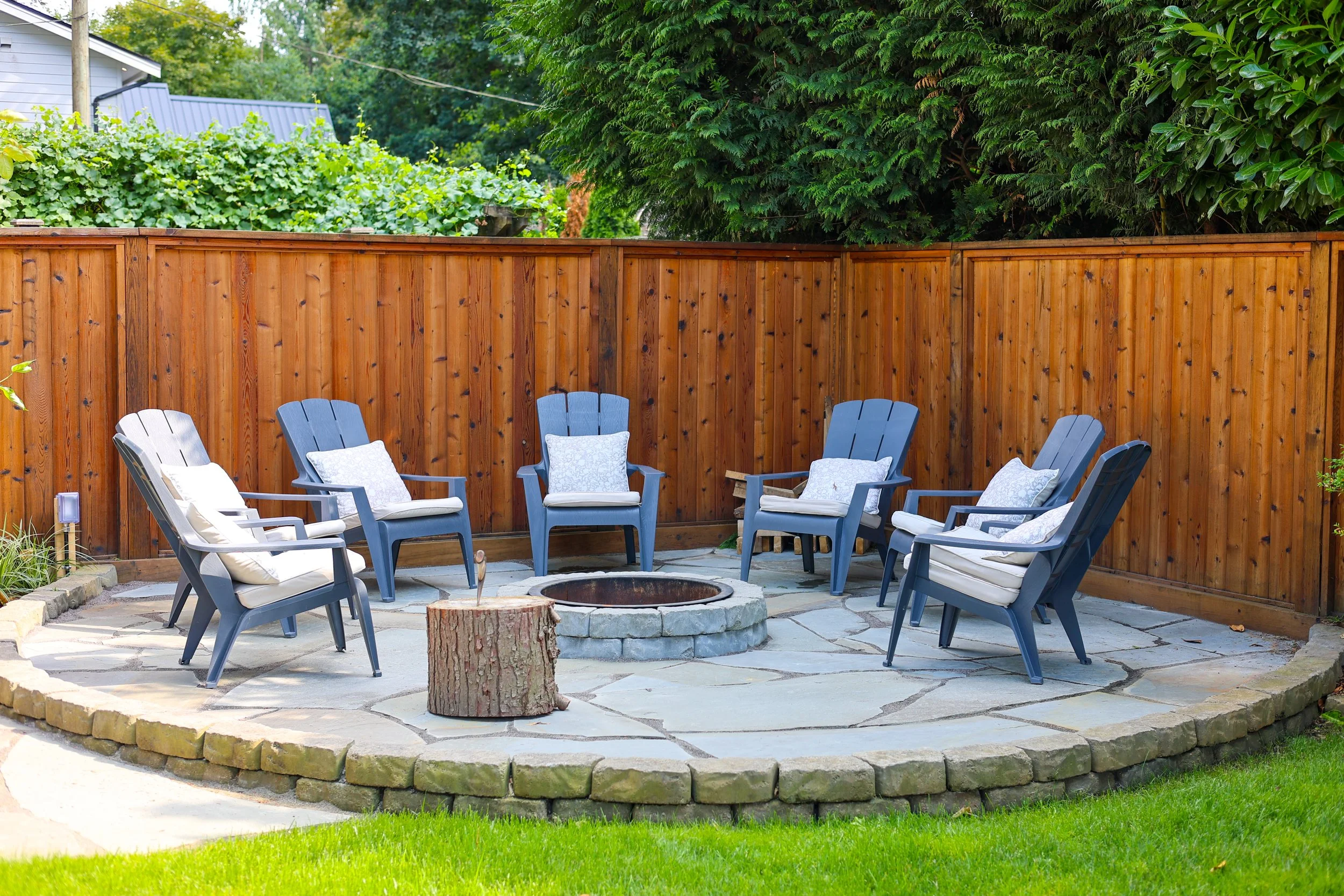 A backyard patio area with six blue Adirondack chairs, each with a white cushion and a decorative pillow, arranged in a circle around a fire pit. There is a wooden fence and lush greenery in the background, including a large tree and some bushes. A t