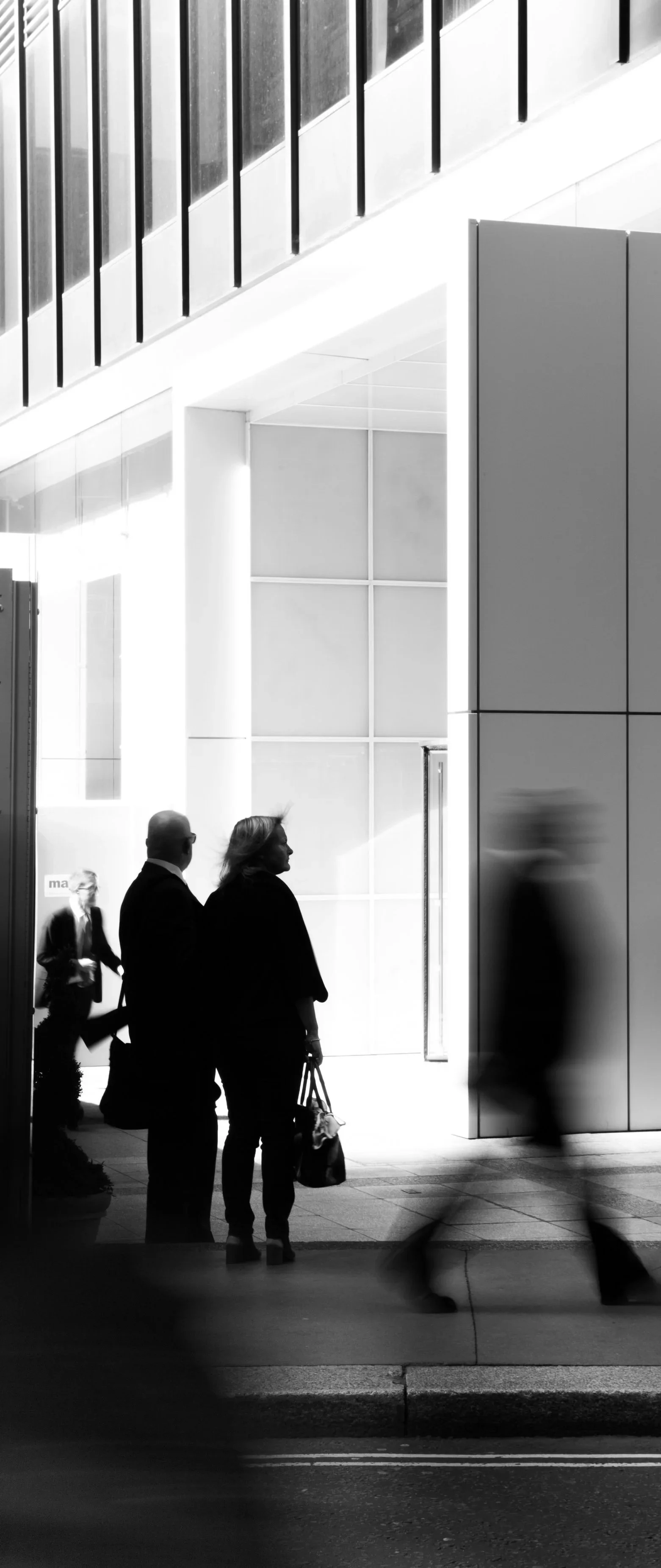 Black and white photo of people walking outside a modern building with large glass windows and sleek architecture, some blurred motion.